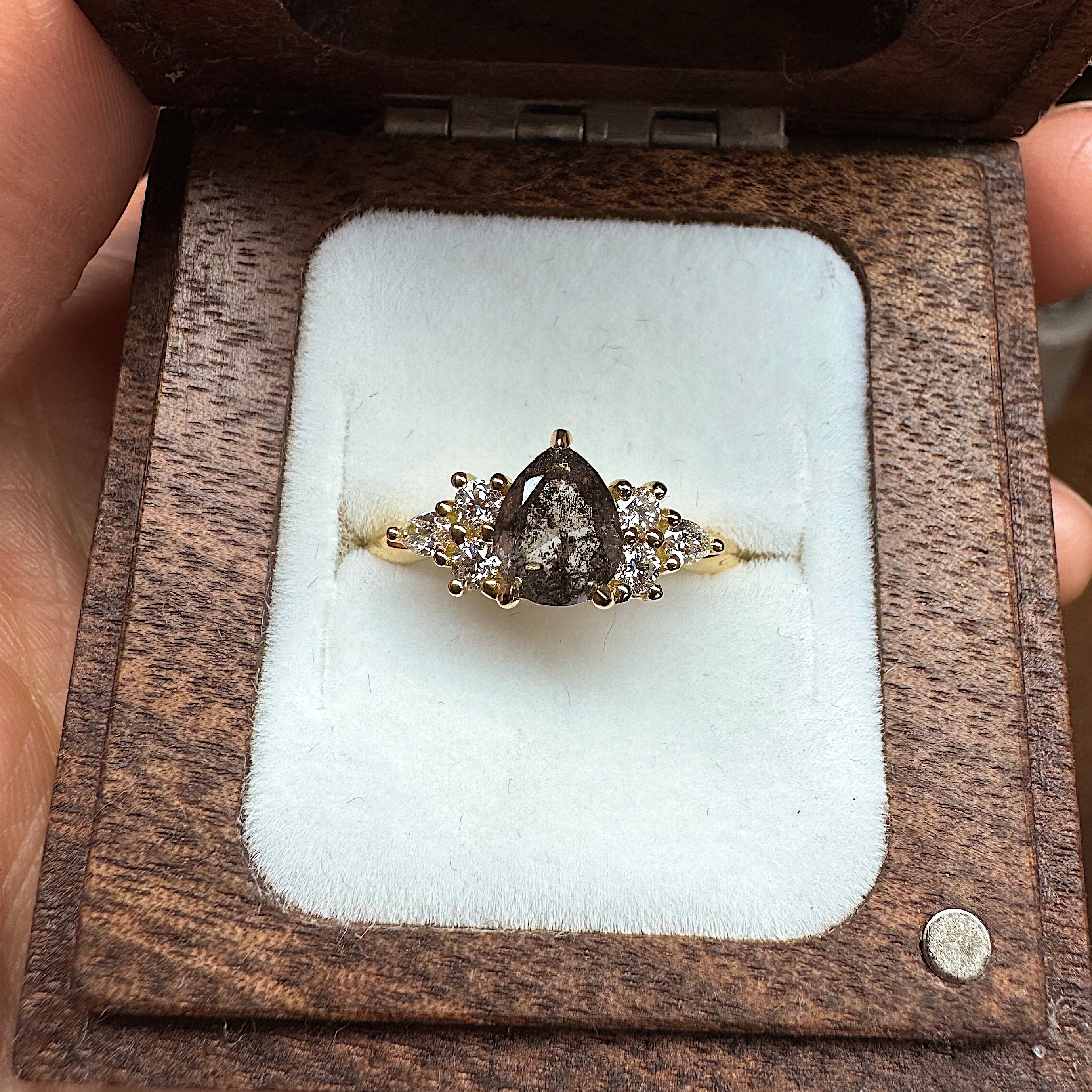 Salt and pepper pear shaped diamond ring in a wooden box held by a hand