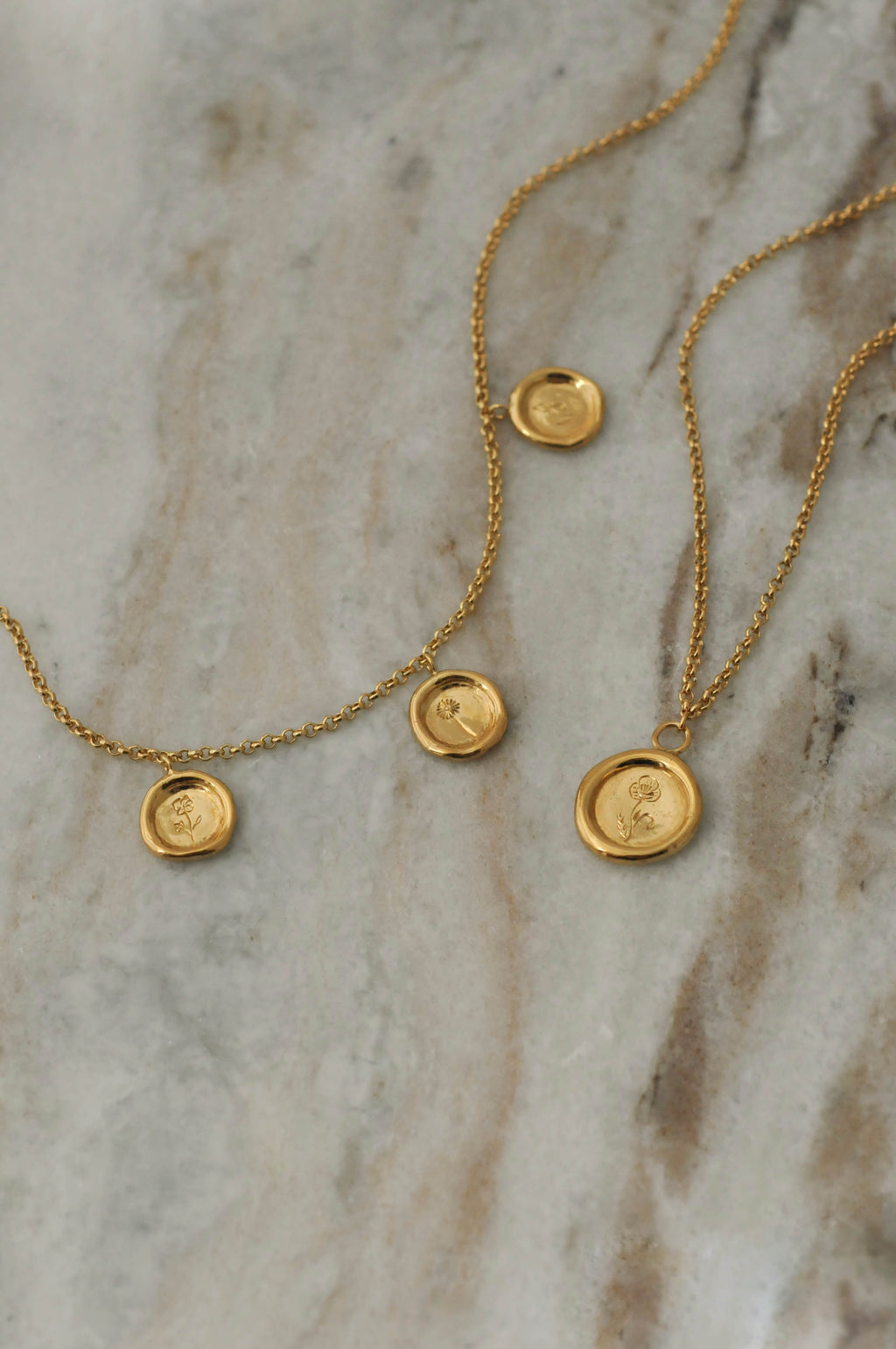 Two delicate gold chain necklaces with small round pendant charms with personal engraved birth flowers, displayed on a light beige marble surface.