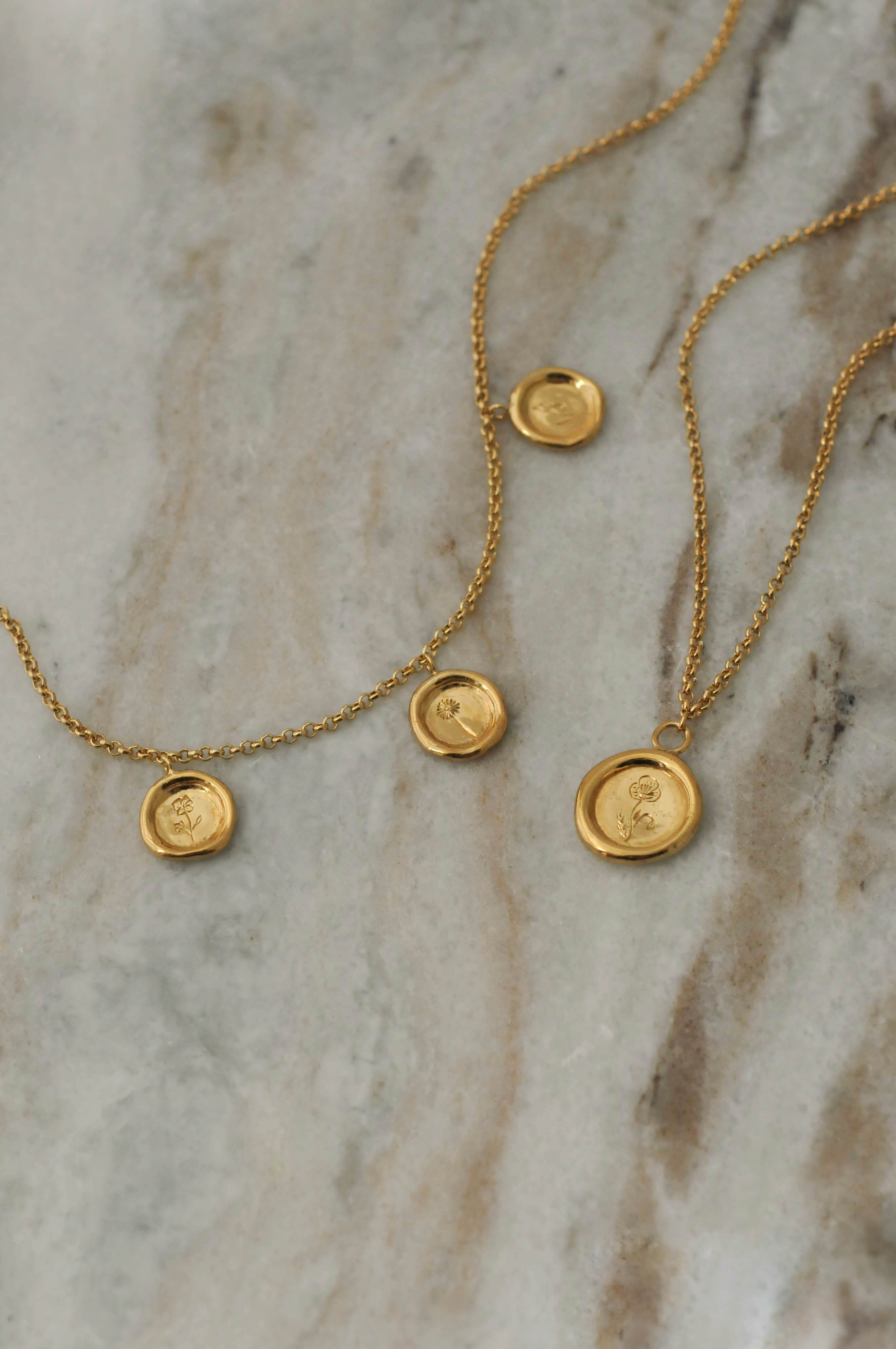 Two delicate gold chain necklaces with small round pendant charms with personal engraved birth flowers, displayed on a light beige marble surface.