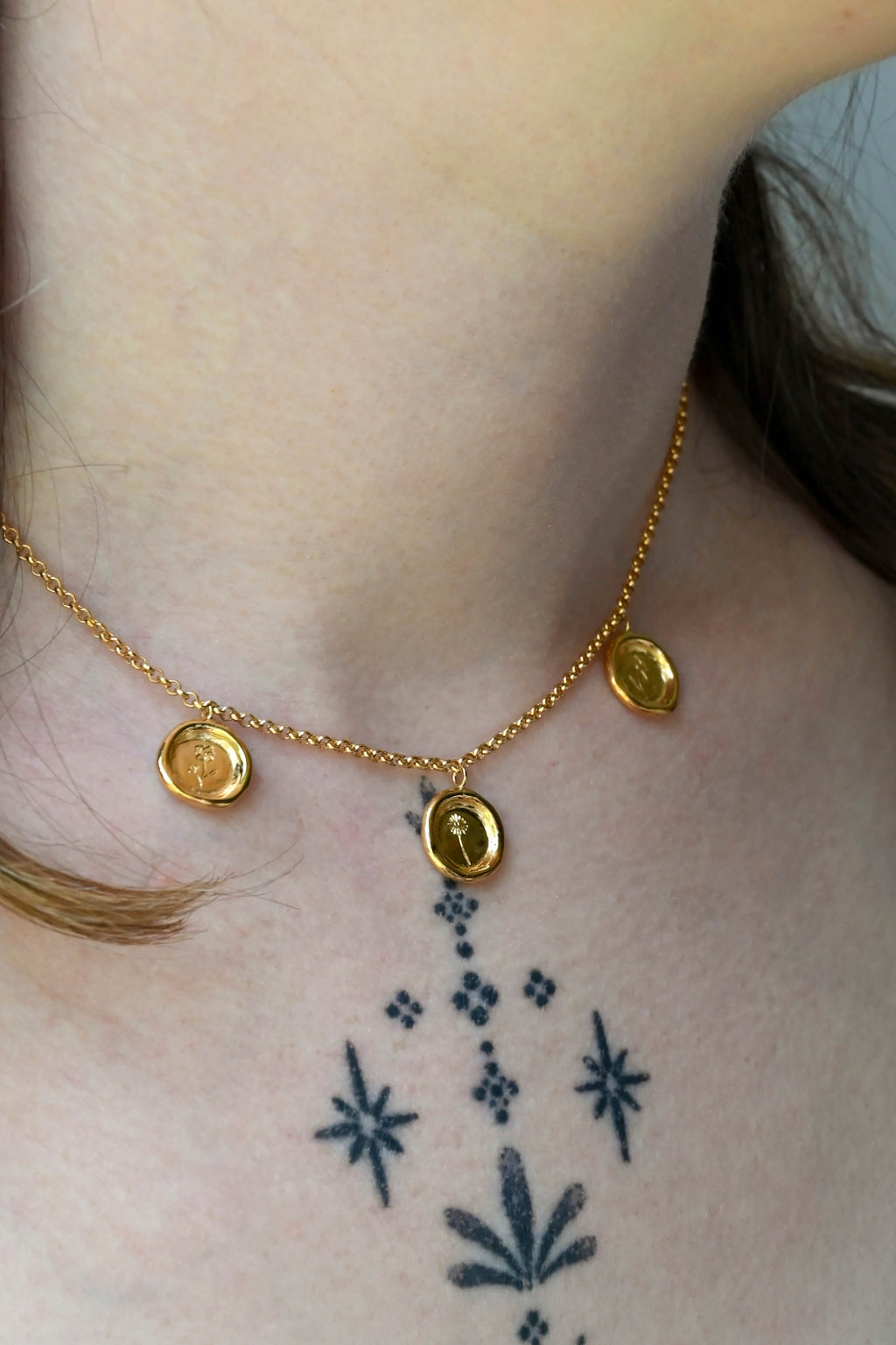 A close-up of a person's neck and upper chest wearing a delicate gold chain necklace with three small circle wax-sealed styled charms with hand engraved flowers . A black floral tattoo is visible on the chest below the necklace.