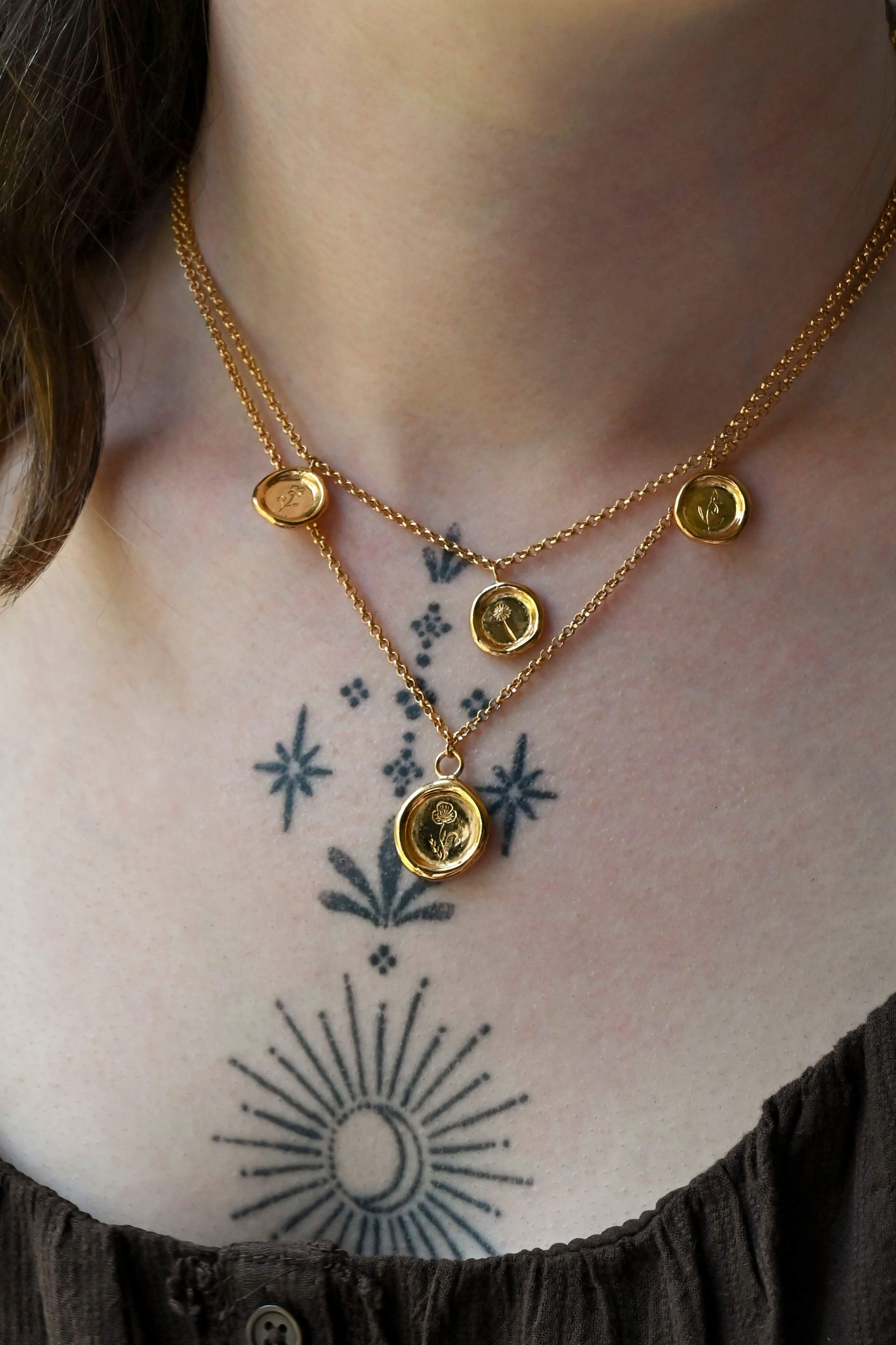 Two layered gold necklaces featuring round wax-seal charm medallions with engraved birth flower motifs, suspended from gold chains against a a woman's neck with tattoos.