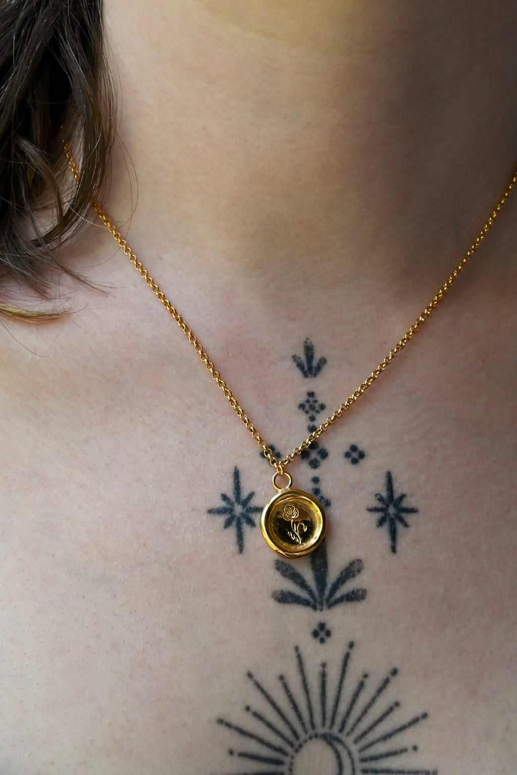 A gold round wax-seal style charm necklace with an engraved birth flower design, suspended from a gold chain against a a woman's neck with tattoos.