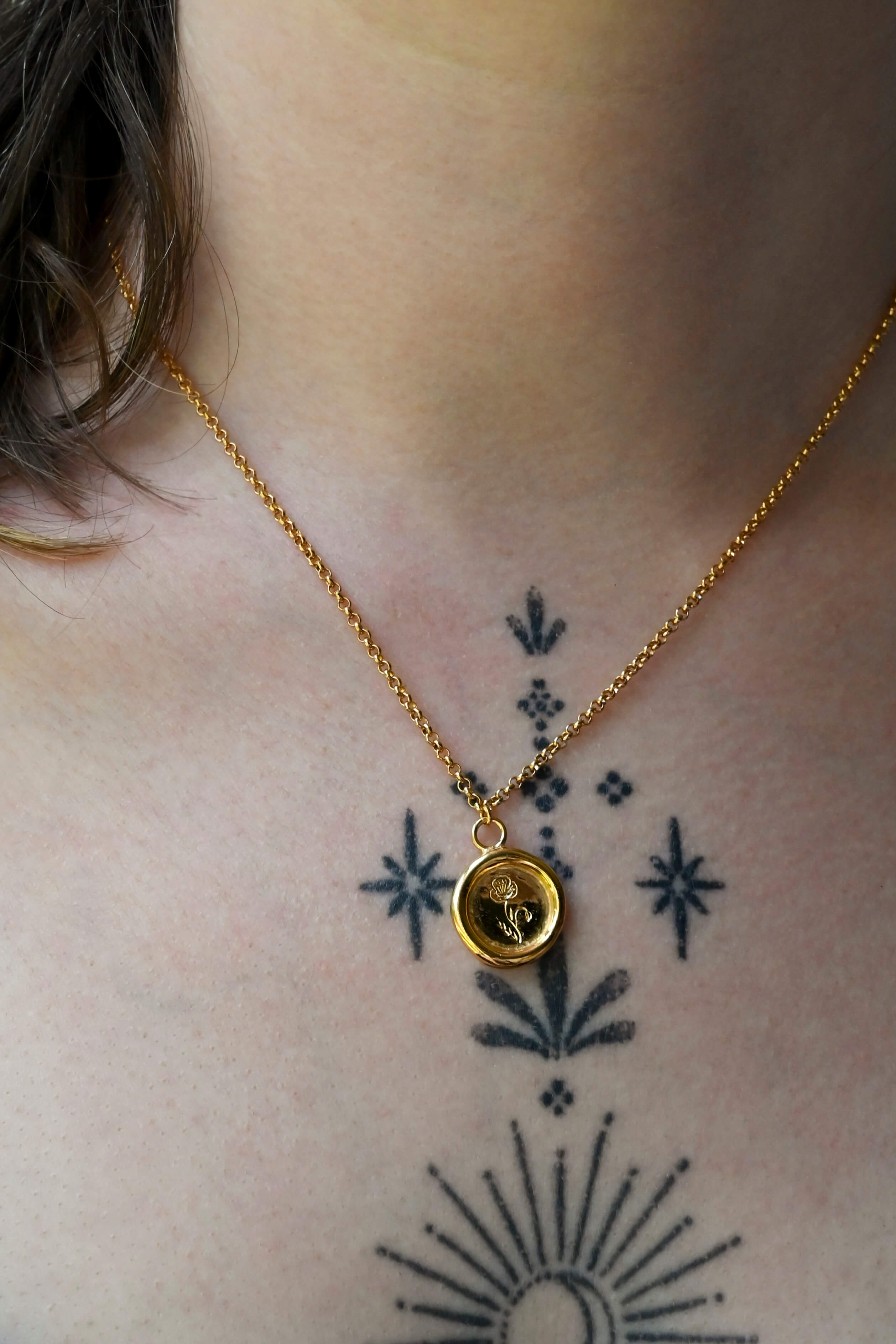 A gold round wax-seal style charm necklace with an engraved birth flower design, suspended from a gold chain against a a woman's neck with tattoos.