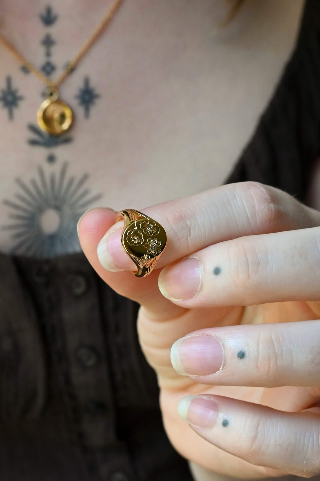 A close-up of a woman's hand holding a small gold signet ring with a circular, textured floral design. The person is wearing a gold necklace and has visible tattoos on their chest and fingers.