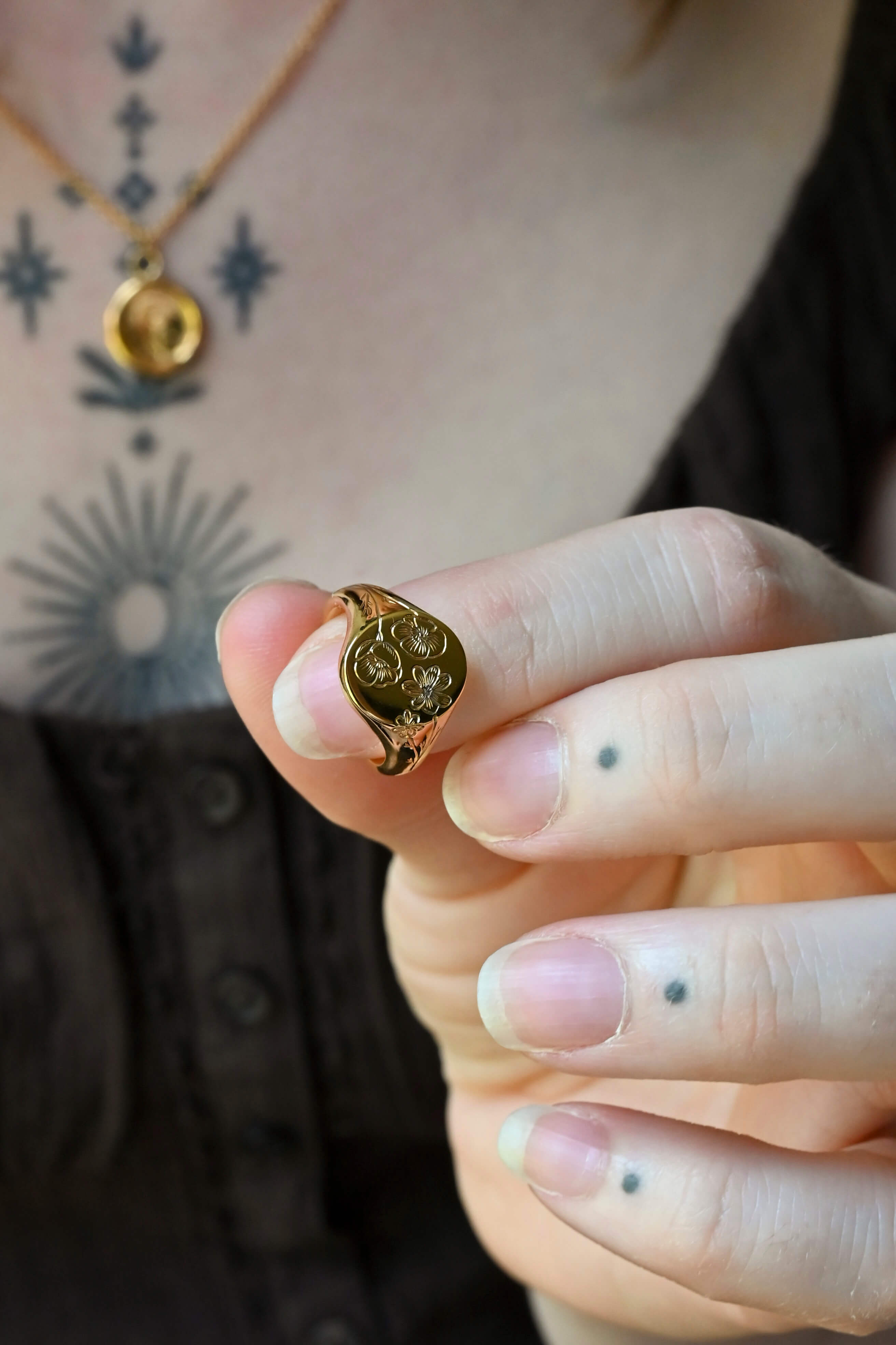 A close-up of a woman's hand holding a small gold signet ring with a circular, textured floral design. The person is wearing a gold necklace and has visible tattoos on their chest and fingers.