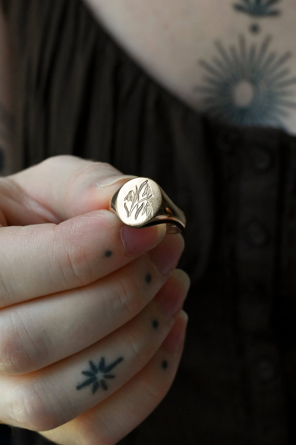 A silver signet ring with an engraved floral birth flower motif. Polished oval pinky signet ring, held by a woman with tattoos.