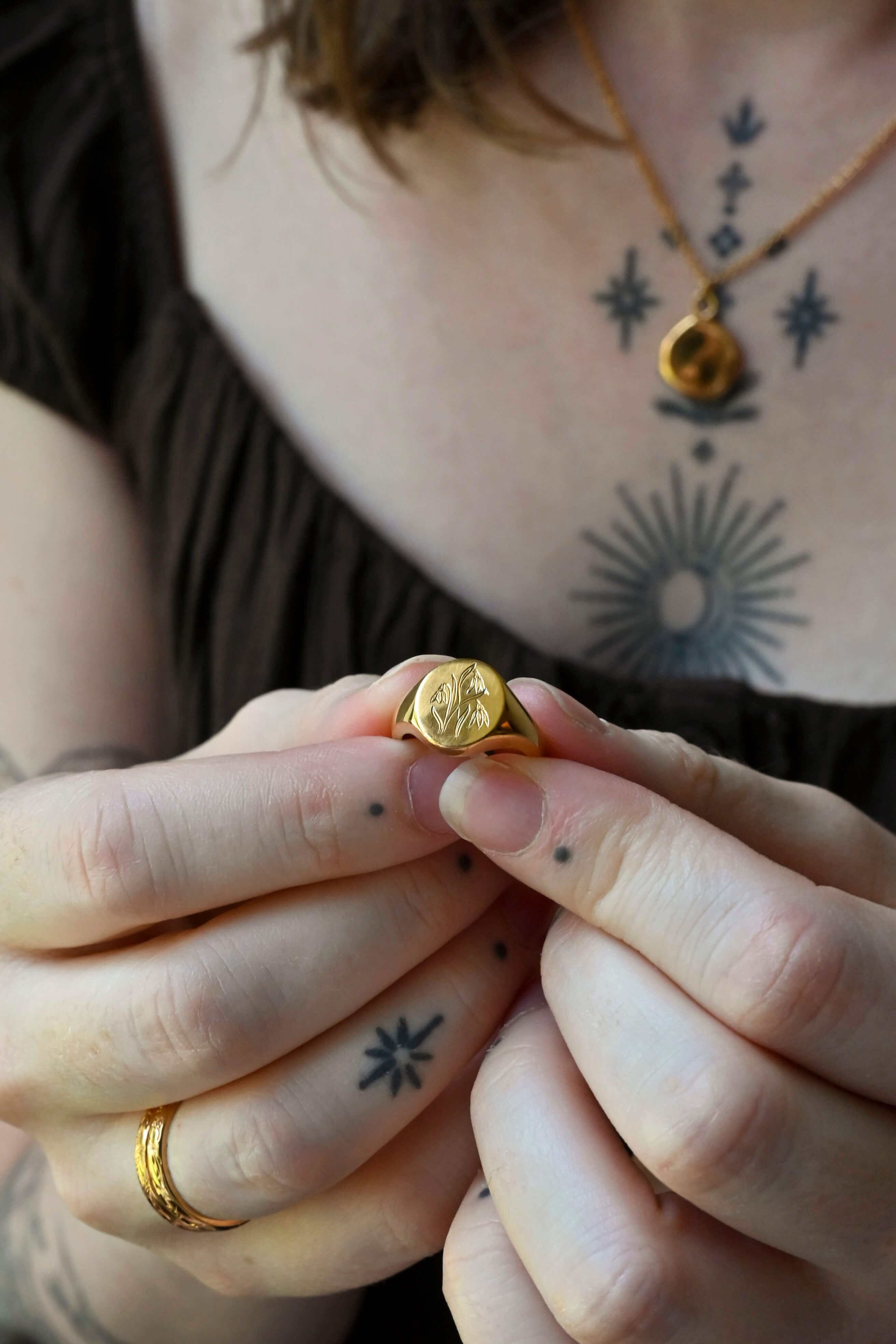 A close-up of a person holding a small gold signet ring with a circular face. They are also wearing a gold necklace and two stacking rings all with unique hand engraved birth flower motifs.