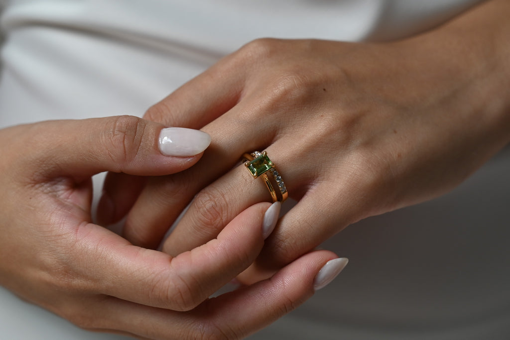 A close-up of a gold ring with a green gemstone being worn on a finger, with another hand gently resting on top. The background shows a soft, out-of-focus white garment.