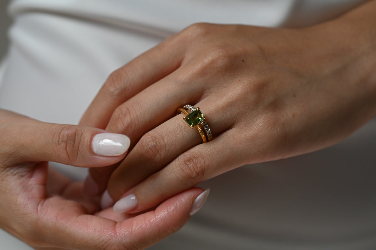 A close-up of a gold ring with a green gemstone worn on a finger, with another hand gently resting on top against a soft white background.