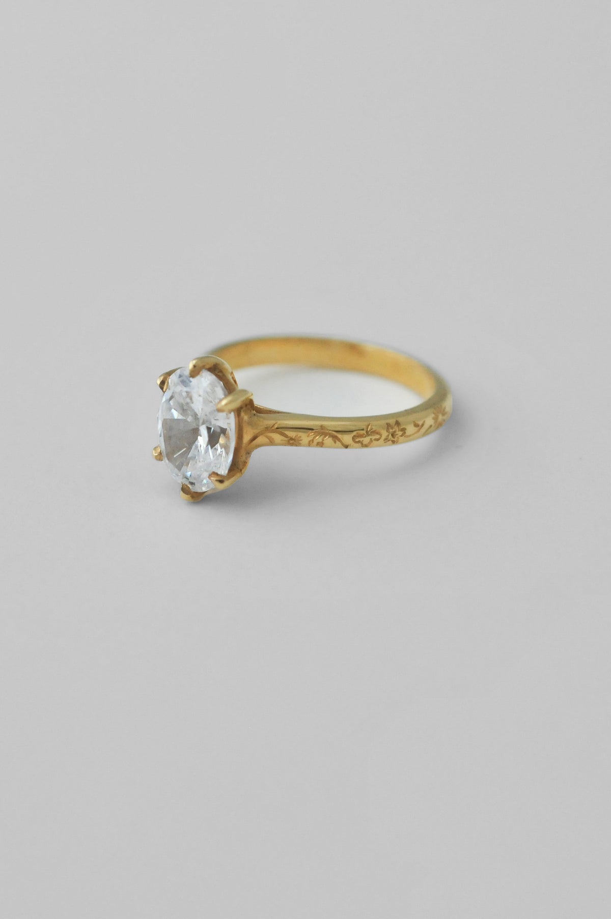 A yellow gold ring with an engraved band and a large oval diamond in a 6prong setting, floral engravings along the band of the ring, photographed on a neutral light gray background.