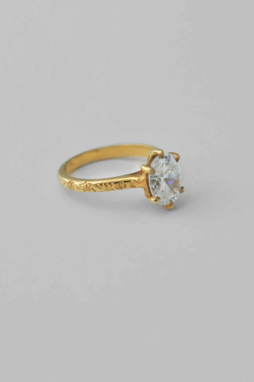 A yellow gold ring with an engraved band and a large oval diamond in a 6prong setting, floral engravings along the band of the ring, photographed on a neutral light gray background.