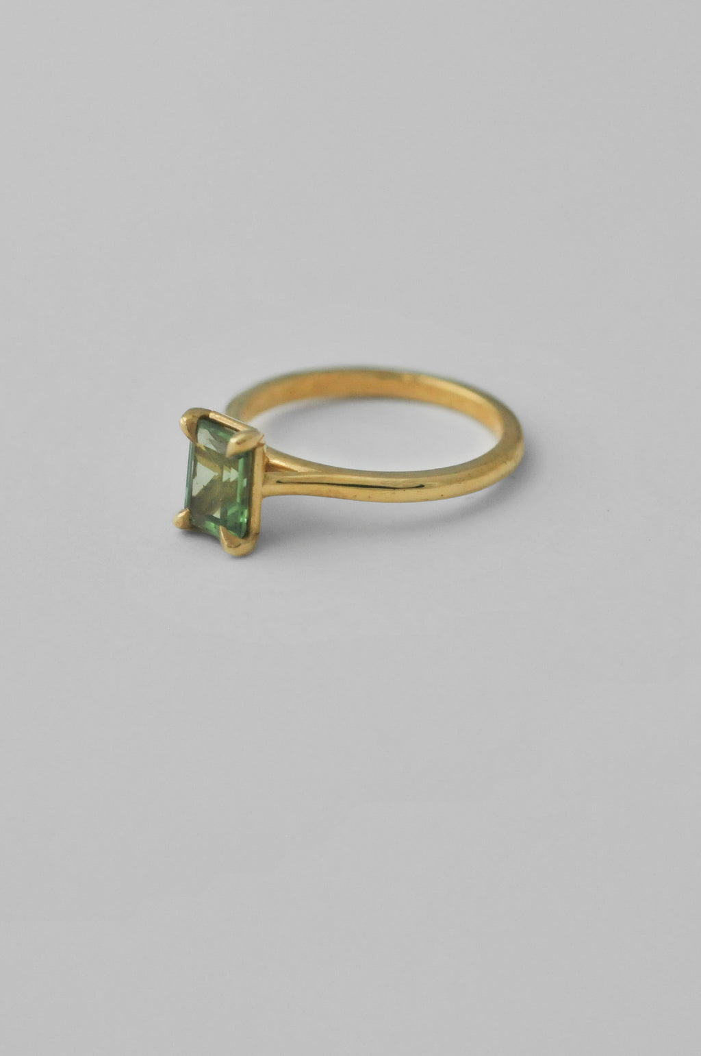 A yellow gold solitaire ring featuring a rectangular emerald-cut green tourmaline gemstone in a four-prong setting, photographed against a neutral light gray background.