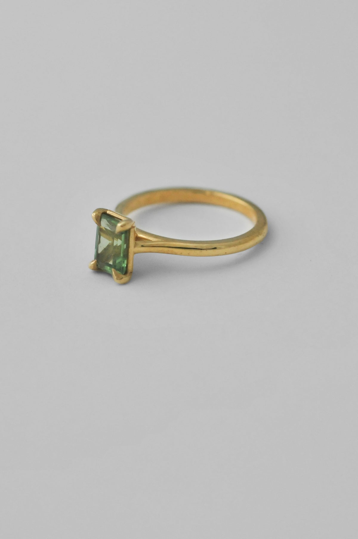 A yellow gold solitaire ring featuring a rectangular emerald-cut green tourmaline gemstone in a four-prong setting, photographed against a neutral light gray background.