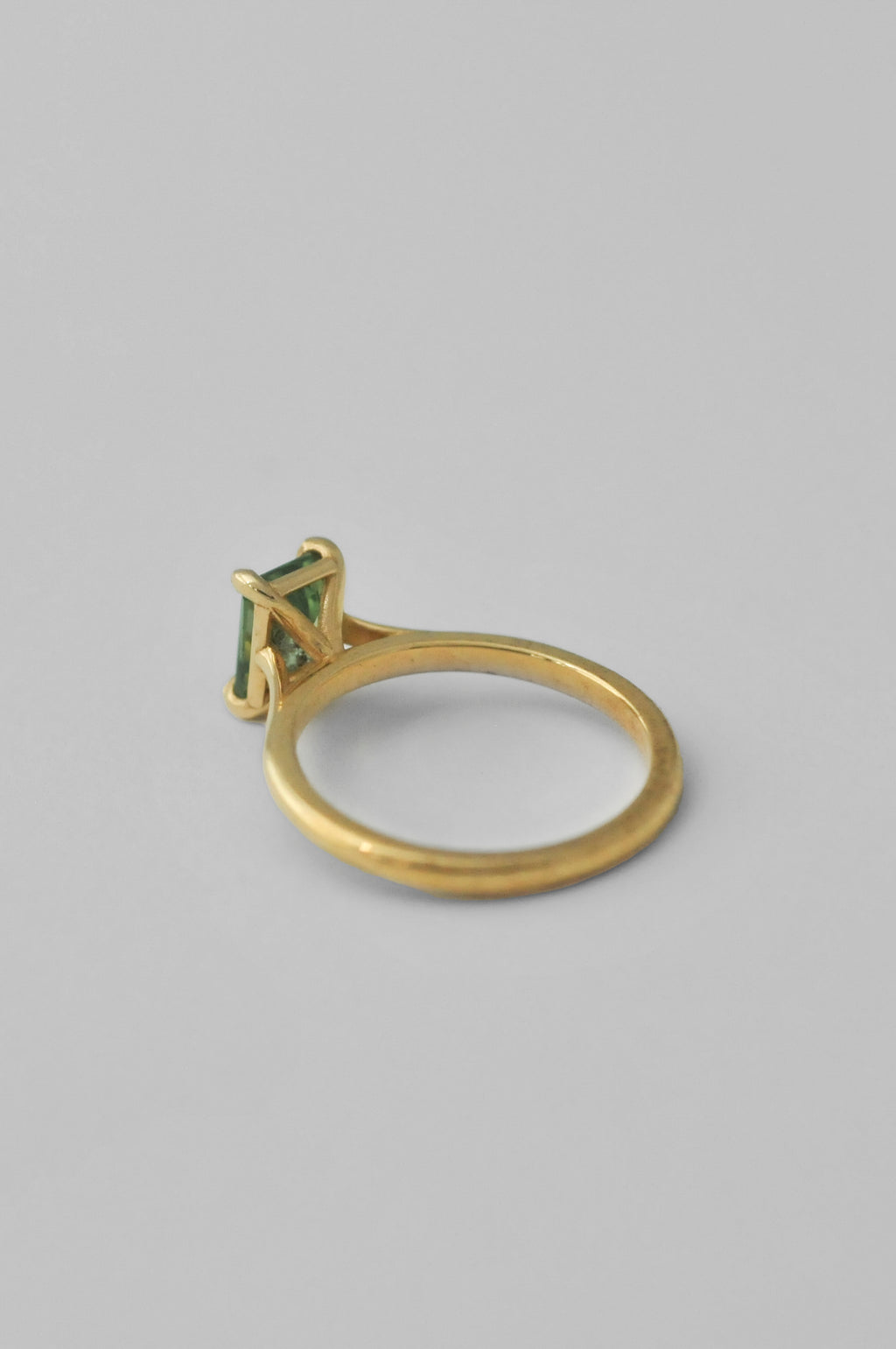 A yellow gold solitaire ring featuring a rectangular emerald-cut green tourmaline gemstone in a four-prong setting, photographed against a neutral light gray background.