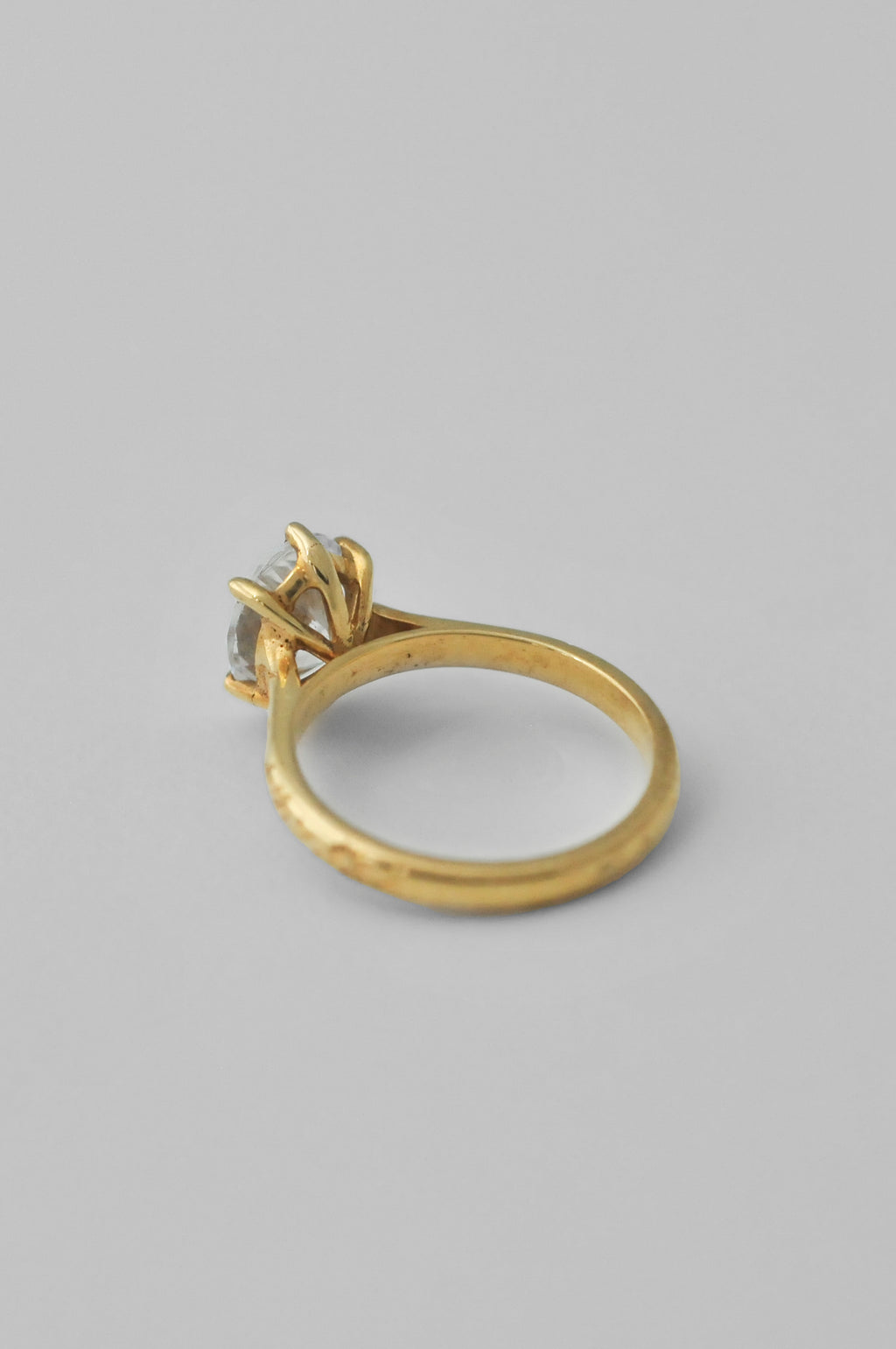 The back of a yellow gold ring with an engraved band and a large oval diamond in a 6 prong setting, floral engravings along the band of the ring, photographed on a neutral light gray background.