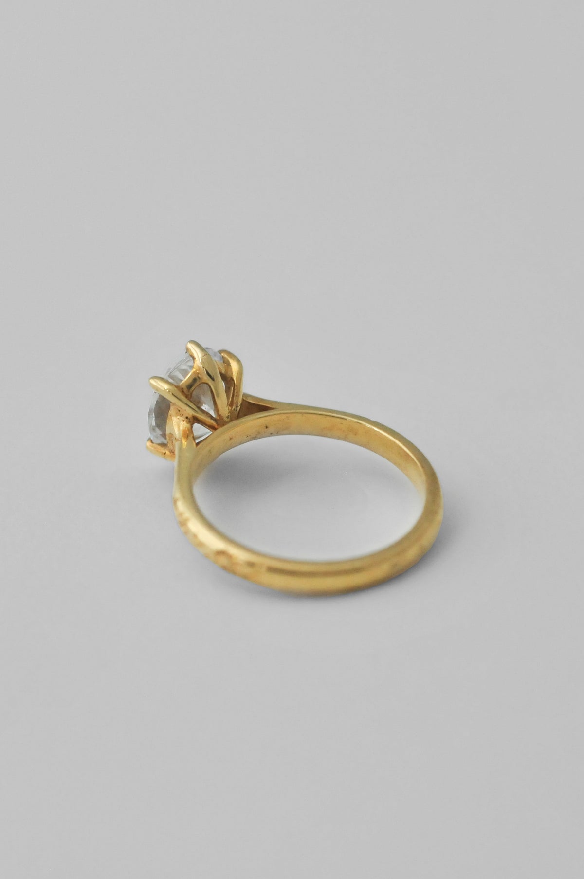 The back of a yellow gold ring with an engraved band and a large oval diamond in a 6 prong setting, floral engravings along the band of the ring, photographed on a neutral light gray background.