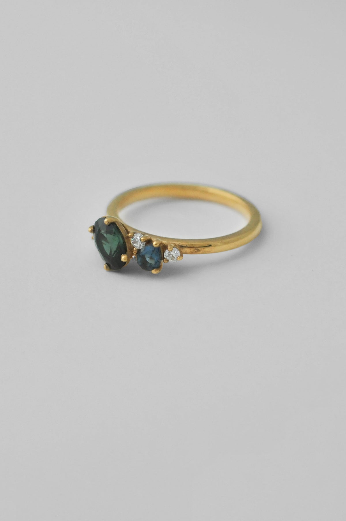 A yellow gold ring featuring a marquise-cut dark green tourmaline gemstone as the centre, flanked by two smaller round topaz gemstones and a small round diamond accent stone, set against a plain light grey background.