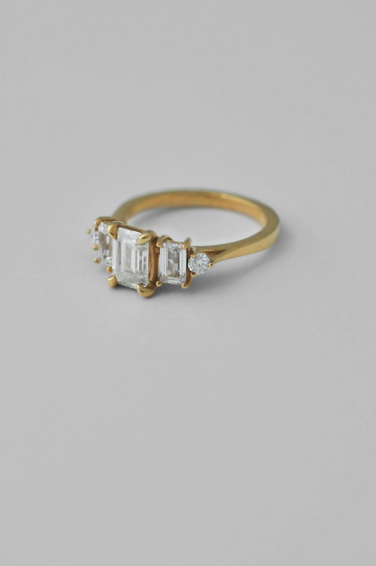 A yellow gold ring featuring a central baguette-cut diamond flanked by two smaller rectangular diamonds and one round diamond, set against a solid light gray background.