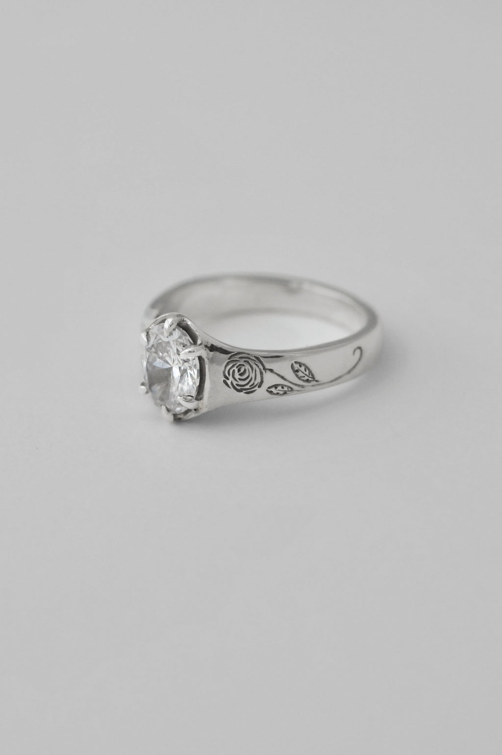 A silver signet ring featuring a large, clear, round cut diamond in a prong setting, with engraved floral roses on the sides of the ring, shown against a neutral light gray background.