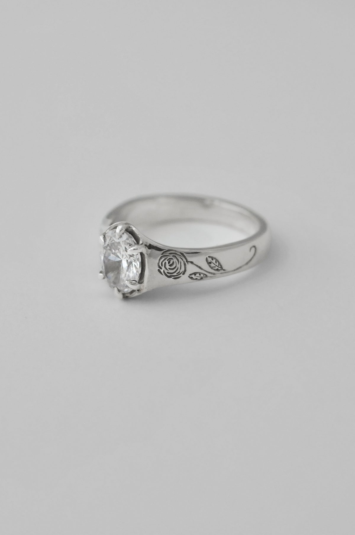 A silver signet ring featuring a large, clear, round cut diamond in a prong setting, with engraved floral roses on the sides of the ring, shown against a neutral light gray background.