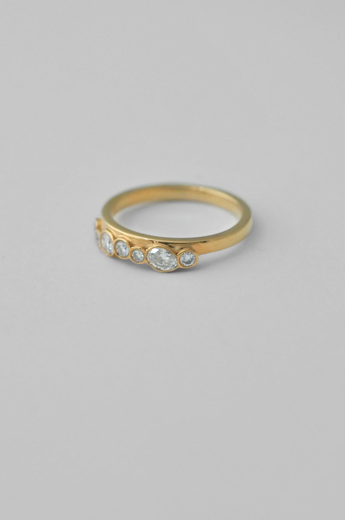 A gold ring with five bezel-set clear oval diamonds, photographed on a neutral light gray background.