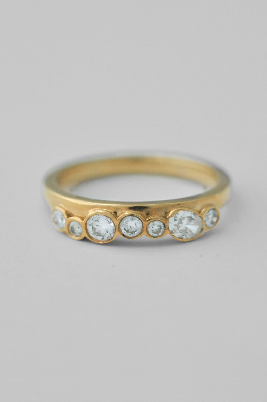 A yellow gold ring featuring a row of bezel-set clear round diamonds of varying sizes, shown on a neutral light gray background.