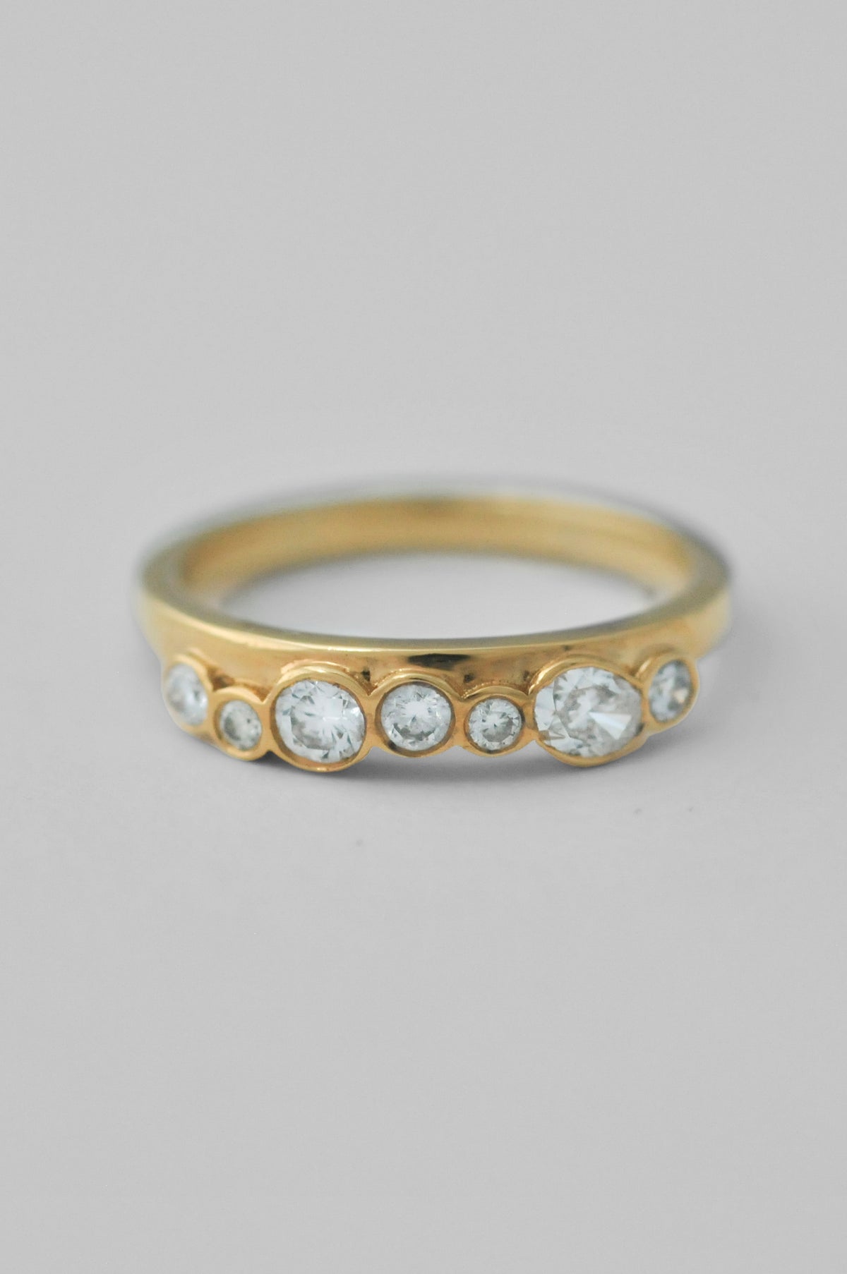 A yellow gold ring featuring a row of bezel-set clear round diamonds of varying sizes, shown on a neutral light gray background.