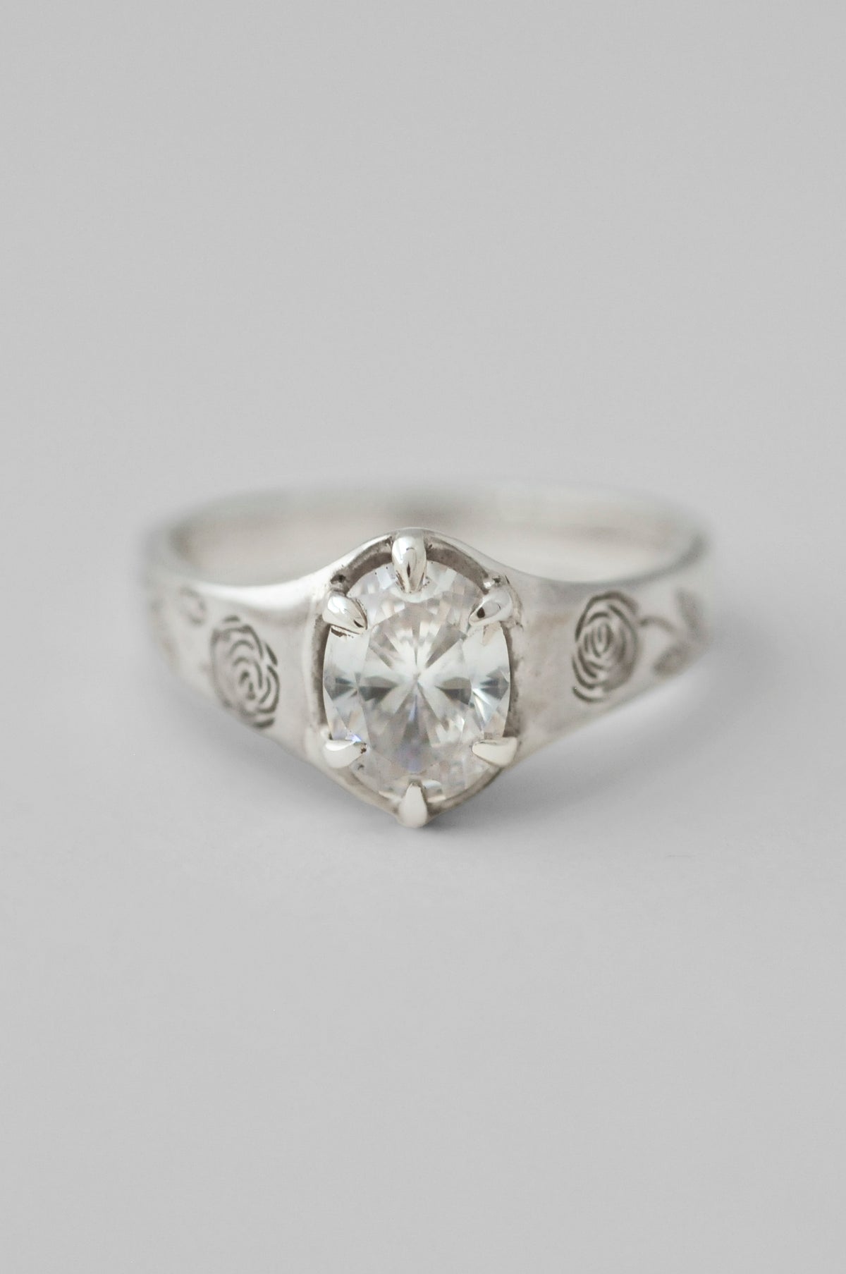 A silver signet ring featuring a large, clear, round cut diamond in a prong setting, with engraved floral roses on the sides of the ring, shown against a neutral light gray background.