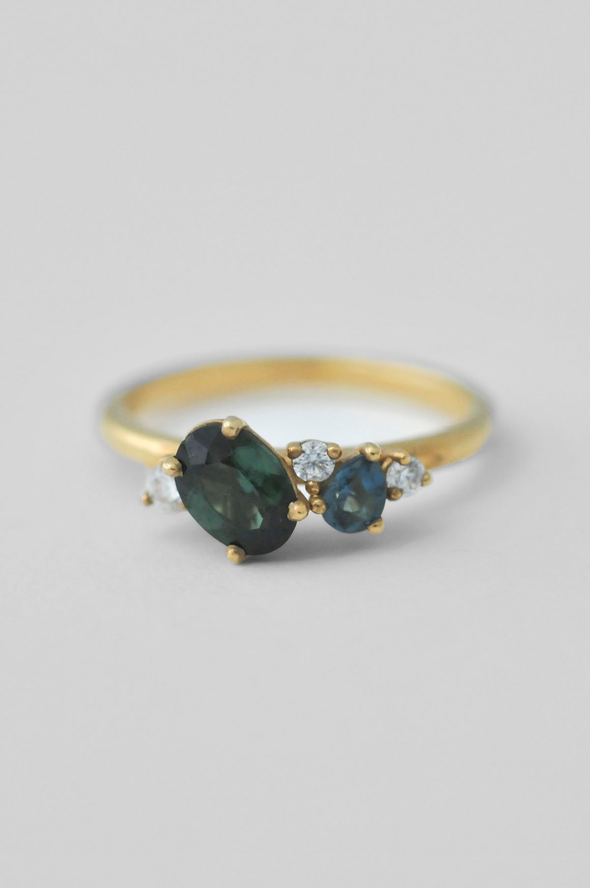 A yellow gold cluster ring featuring a large oval dark green tourmaline gemstone, a smaller oval deep blue topaz gemstone, and two round diamond accent stones, set against a neutral light gray background.