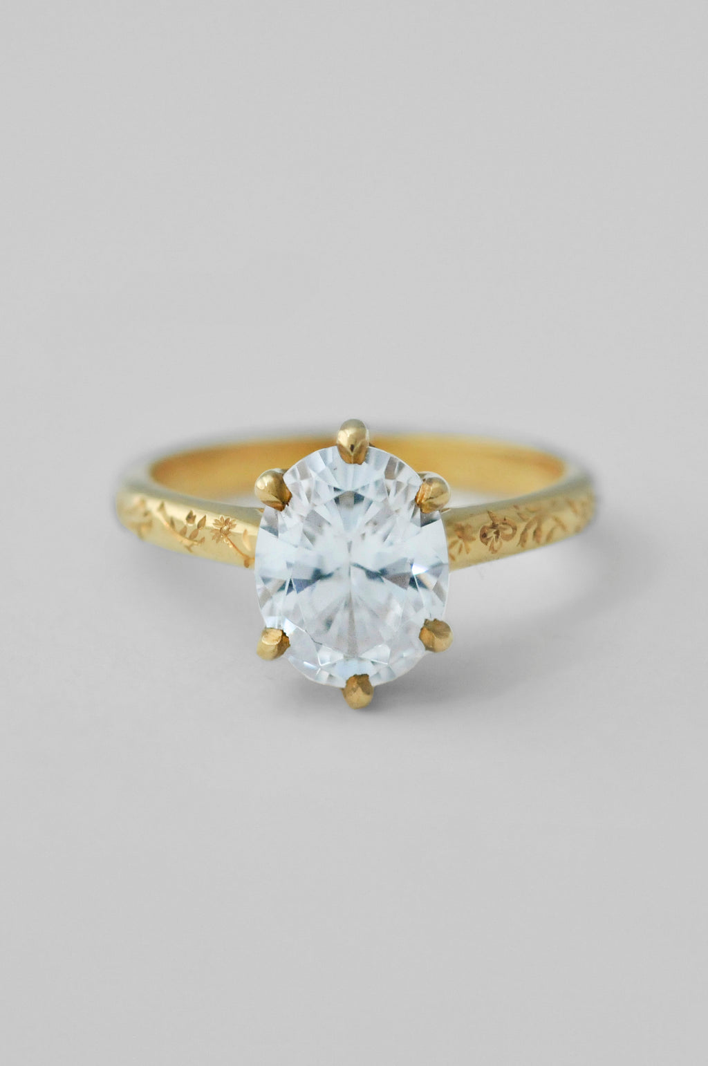 A yellow gold ring with an engraved band and a large oval diamond in a 6prong setting, floral engravings along the band of the ring, photographed on a neutral light gray background.