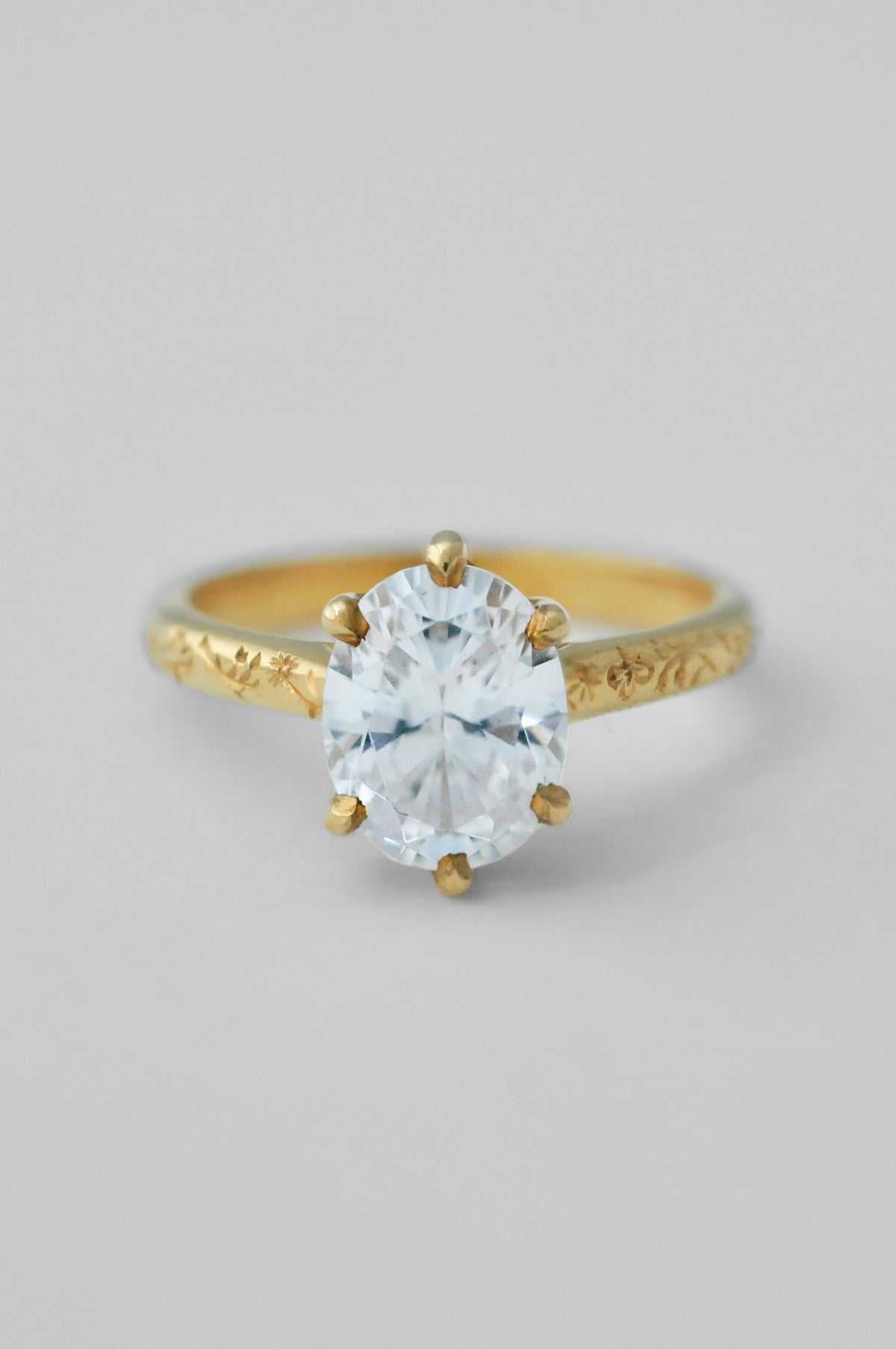 A yellow gold ring with an engraved band and a large oval diamond in a 6prong setting, floral engravings along the band of the ring, photographed on a neutral light gray background.