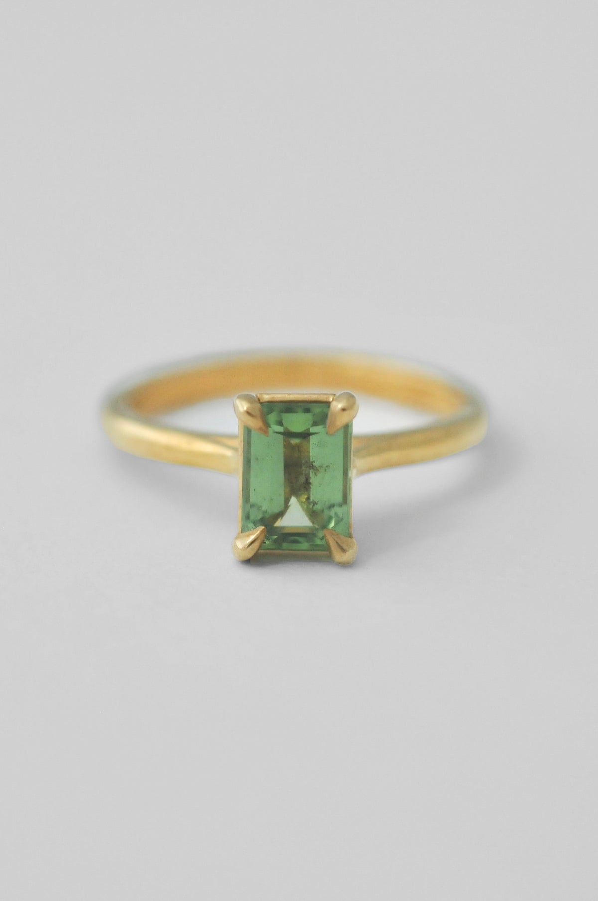 A yellow gold solitaire ring featuring a rectangular emerald-cut green tourmaline gemstone in a four-prong setting, photographed against a neutral light gray background.