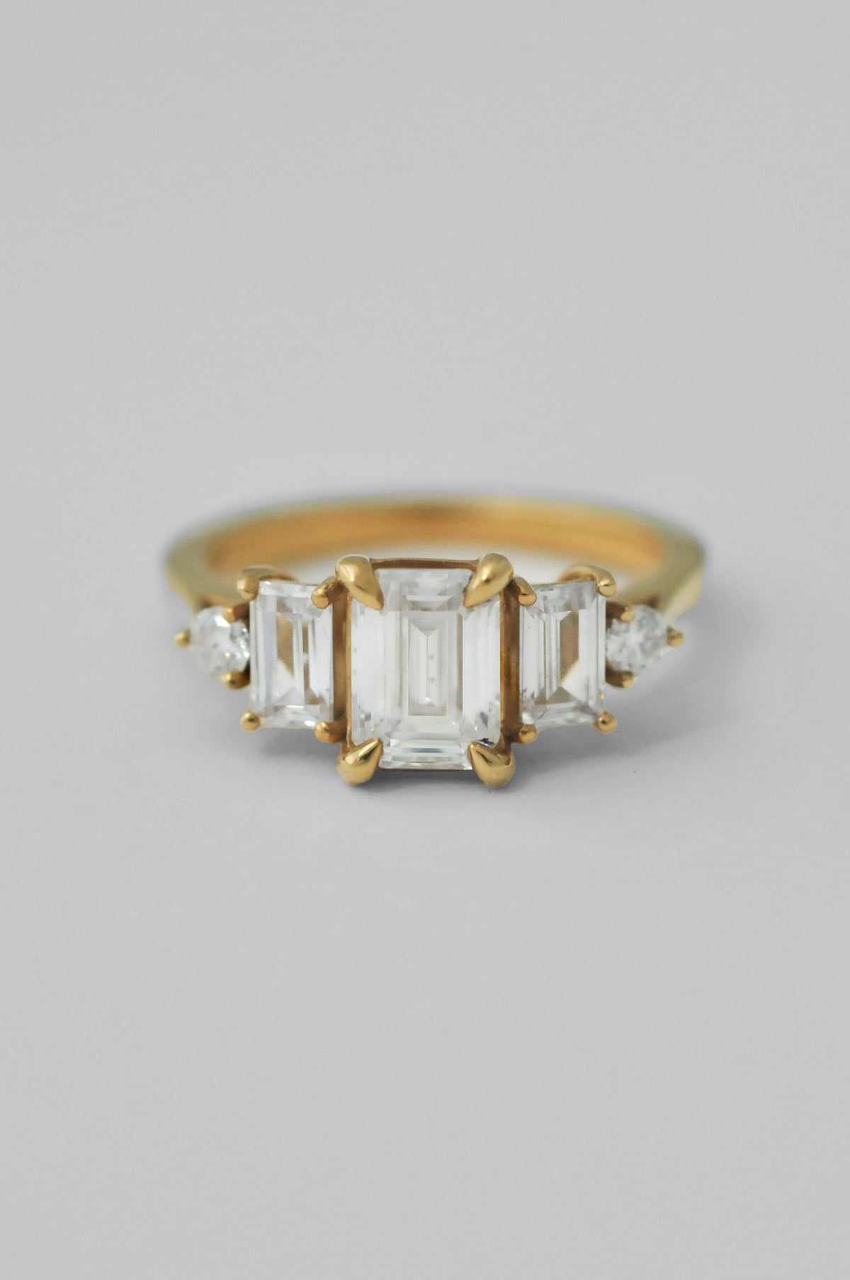 A yellow gold ring featuring a three-stone emerald-cut diamond center with smaller round diamonds on either side, set against a neutral light gray background.