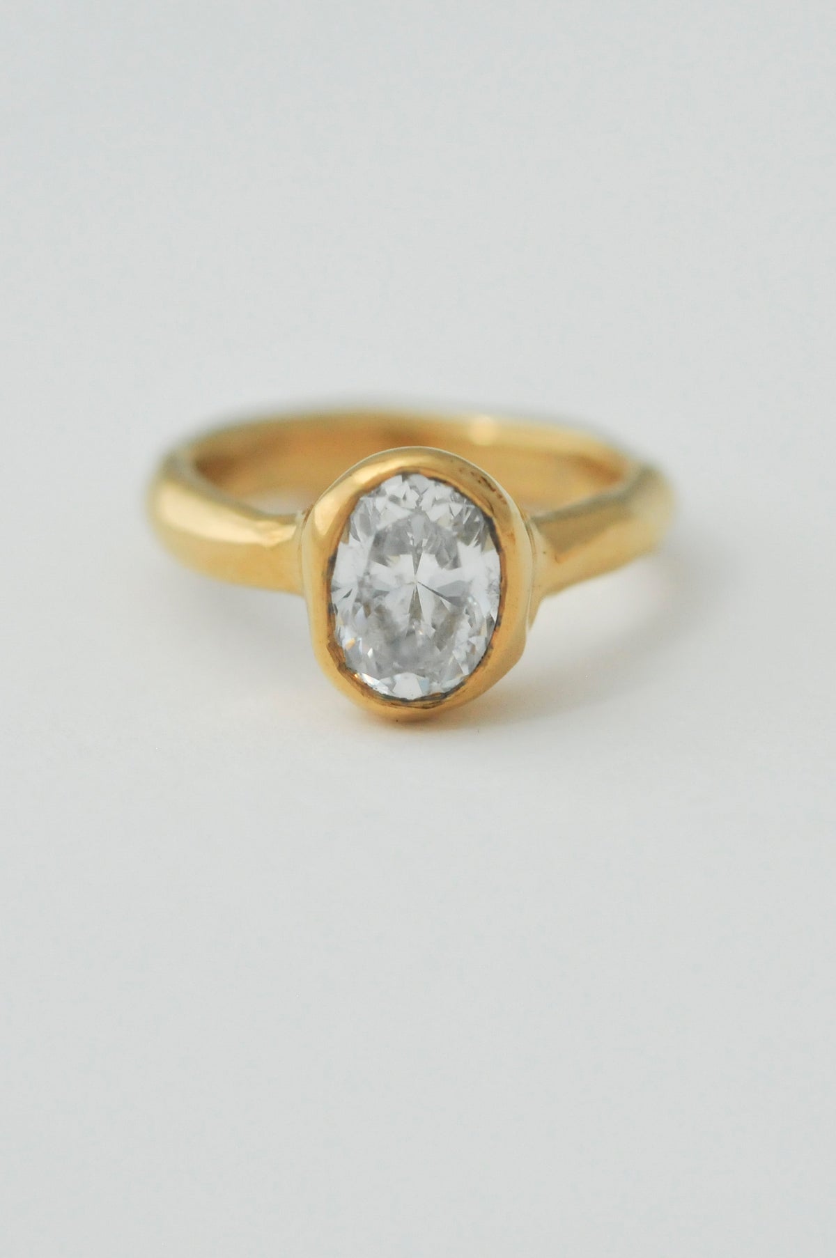 A yellow gold ring featuring an oval-cut diamond in a bezel setting, molten set ring photographed on a clean, light gray background.