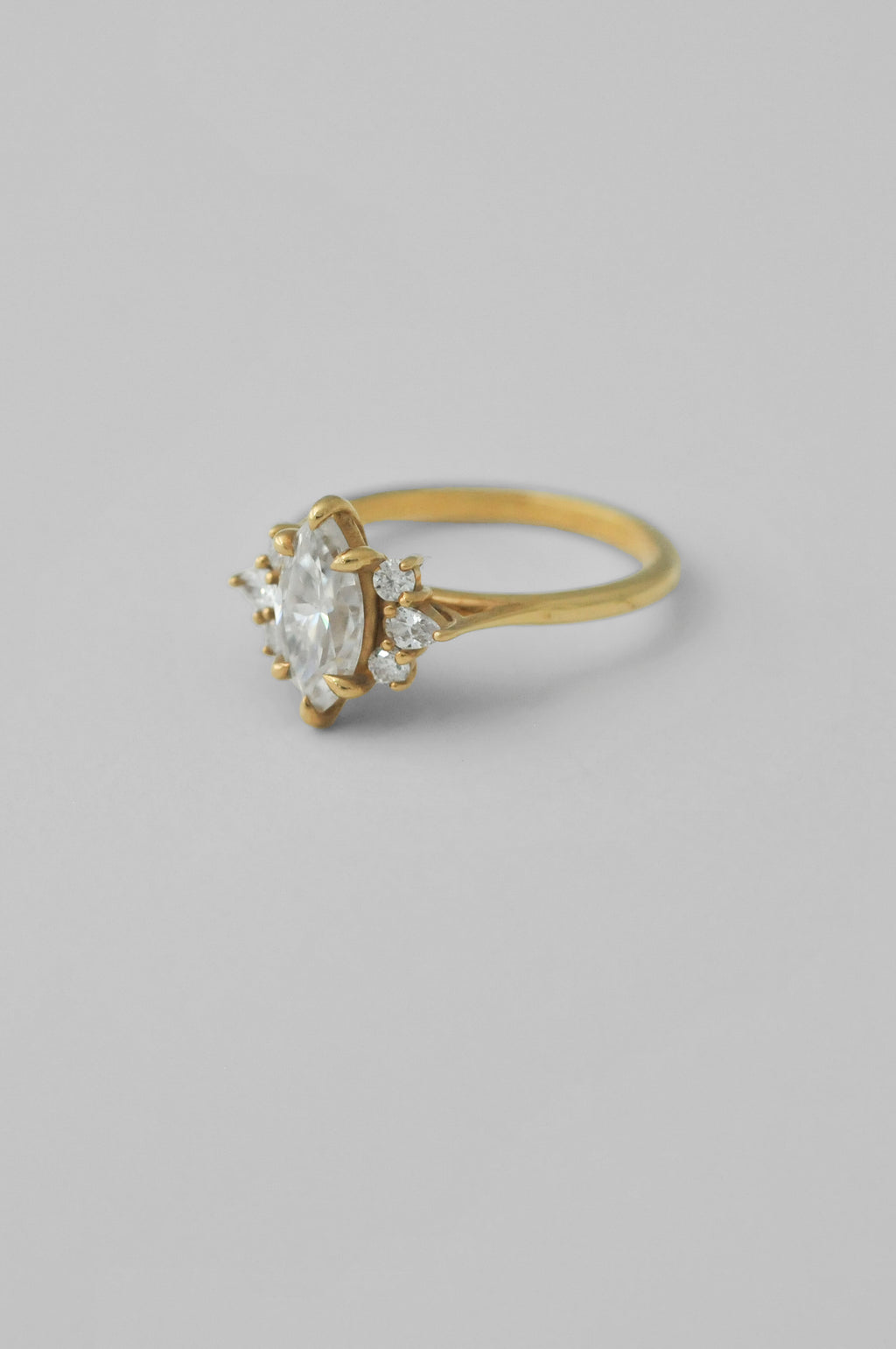 A yellow gold ring featuring a large oval-cut clear diamond surrounded by small accent diamonds, set on a plain light gray background.