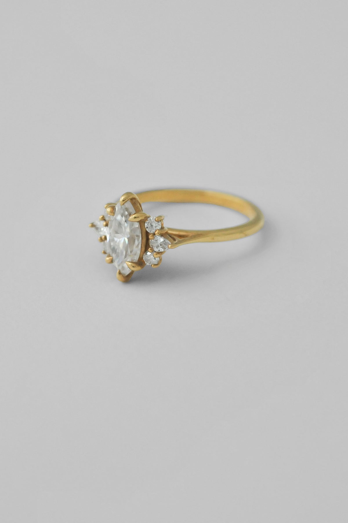 A yellow gold ring featuring a large oval-cut clear diamond surrounded by small accent diamonds, set on a plain light gray background.