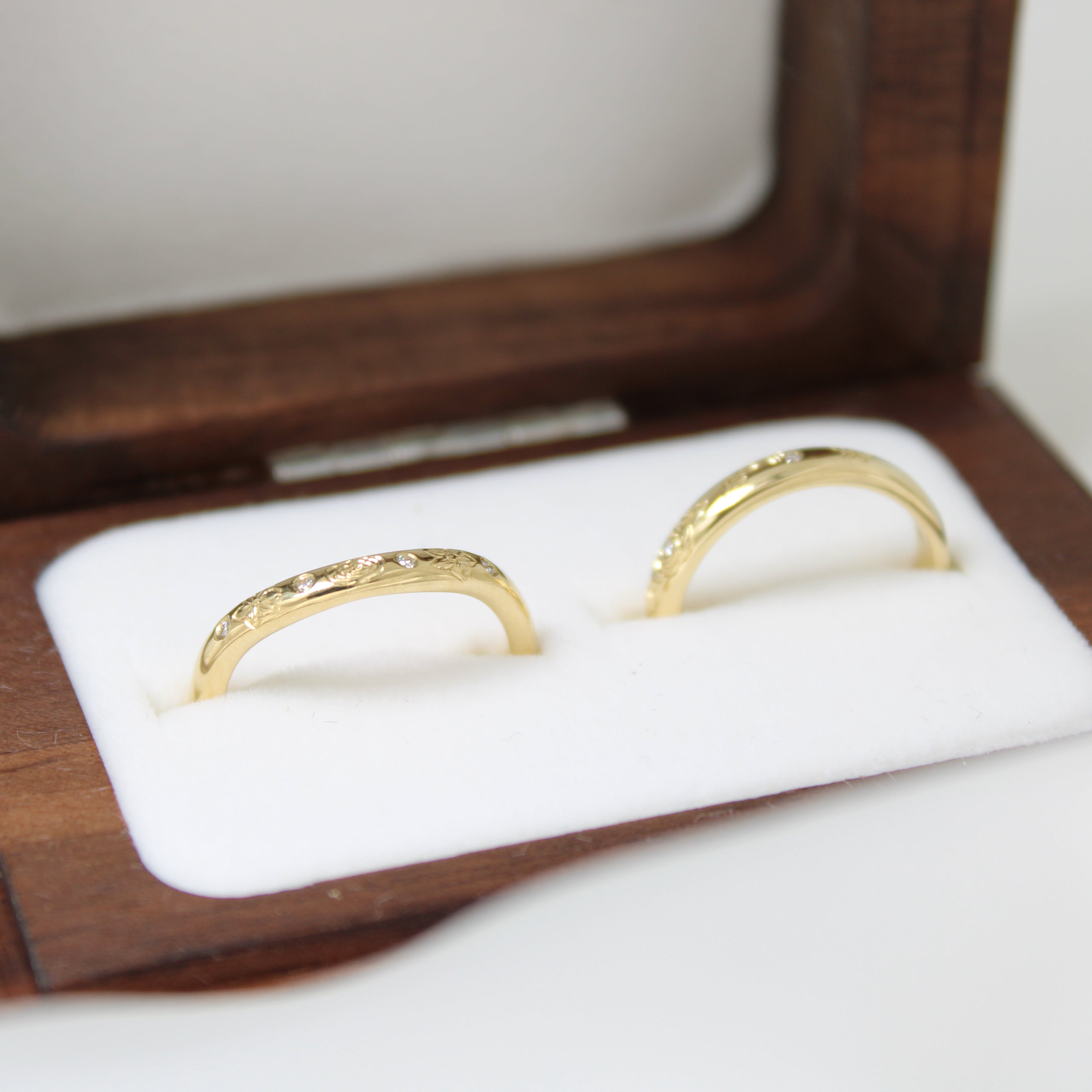 Two gold wedding rings with intricate engraving and diamonds in a wooden box on a white background