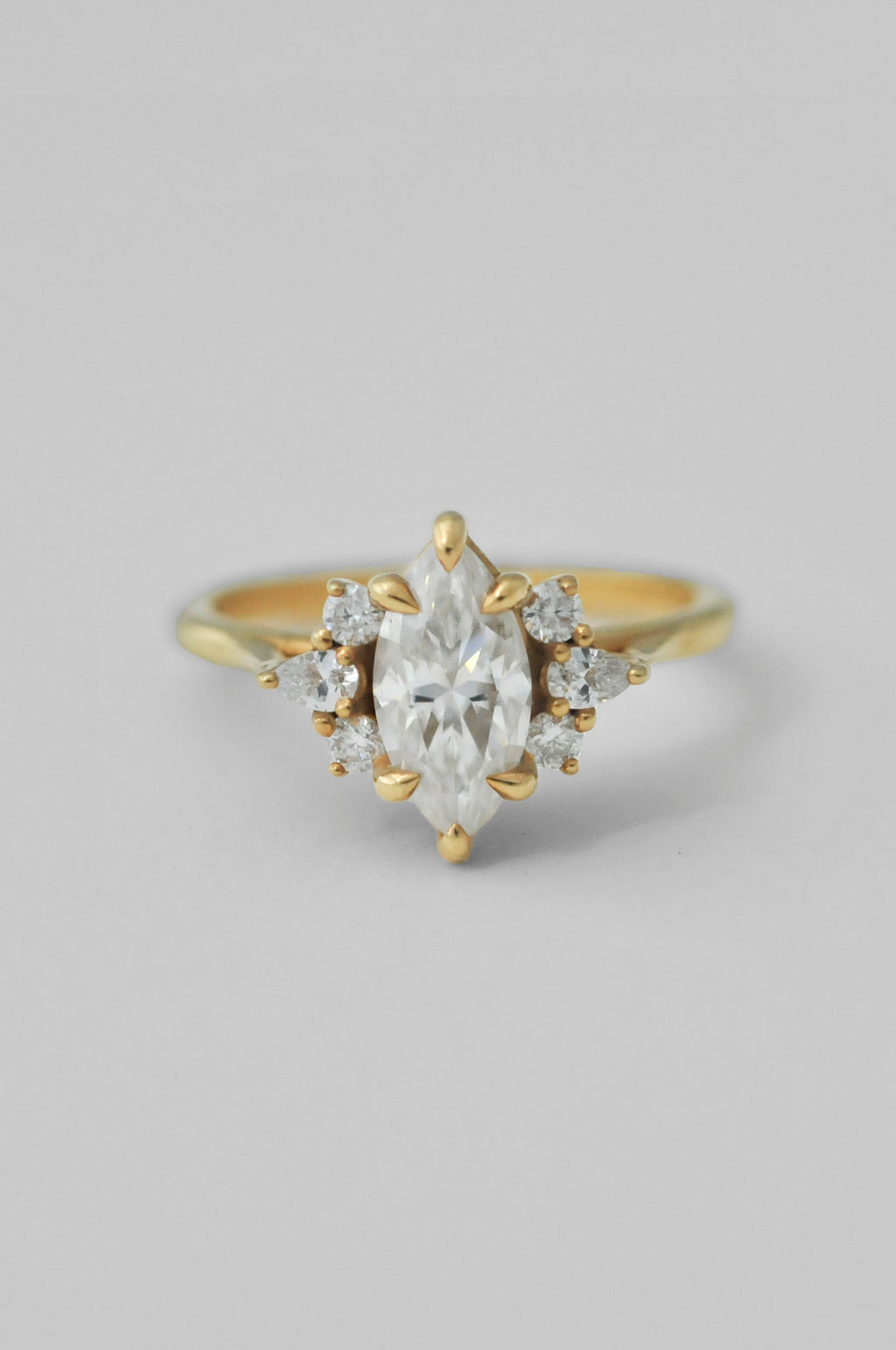 A yellow gold ring featuring a marquise-cut clear center diamond surrounded by small round diamonds on either side, set against a plain light gray background.