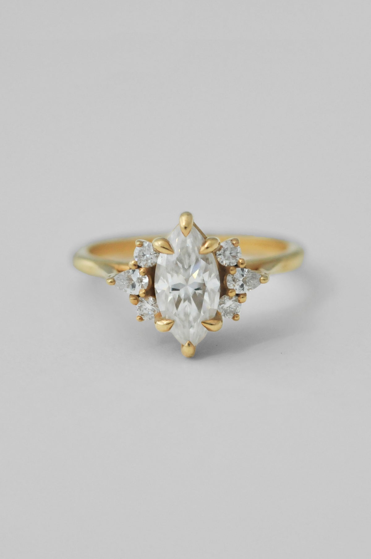 A yellow gold ring featuring a marquise-cut clear center diamond surrounded by small round diamonds on either side, set against a plain light gray background.
