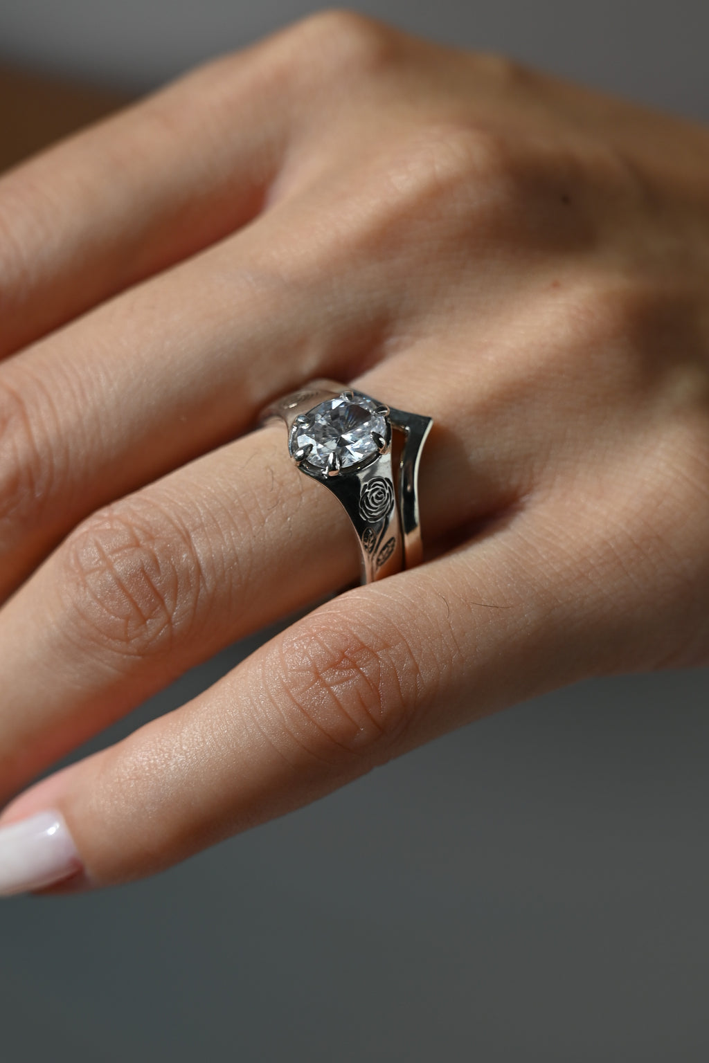 A close-up of a hand wearing a silver ring with a large clear round gemstone and engraved details on the band, set against a soft gray background.