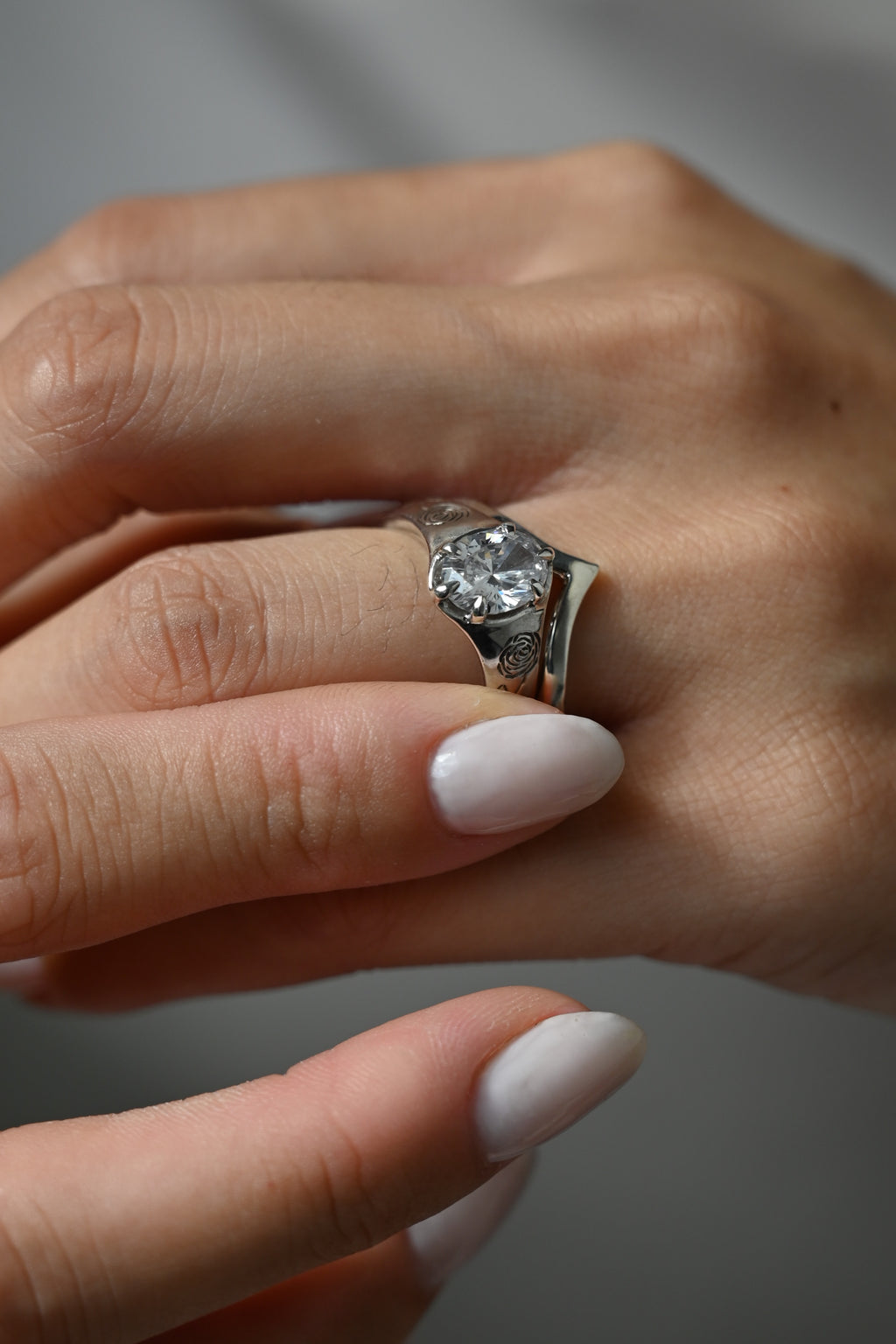 A close-up of a hand wearing a silver-toned ring with a large round clear gemstone. The ring is shown on the middle finger against a soft gray background.