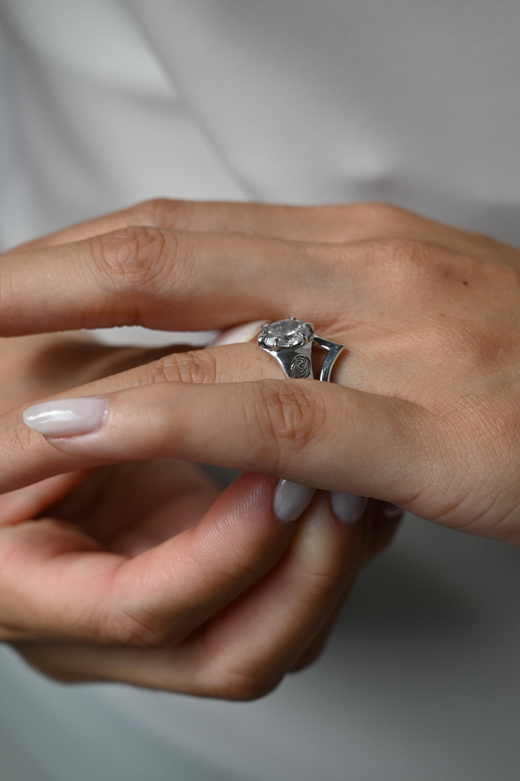 A close-up of a person's hand wearing a silver-toned ring with a large round clear gemstone and side rose flower engravings, against a soft light gray background.