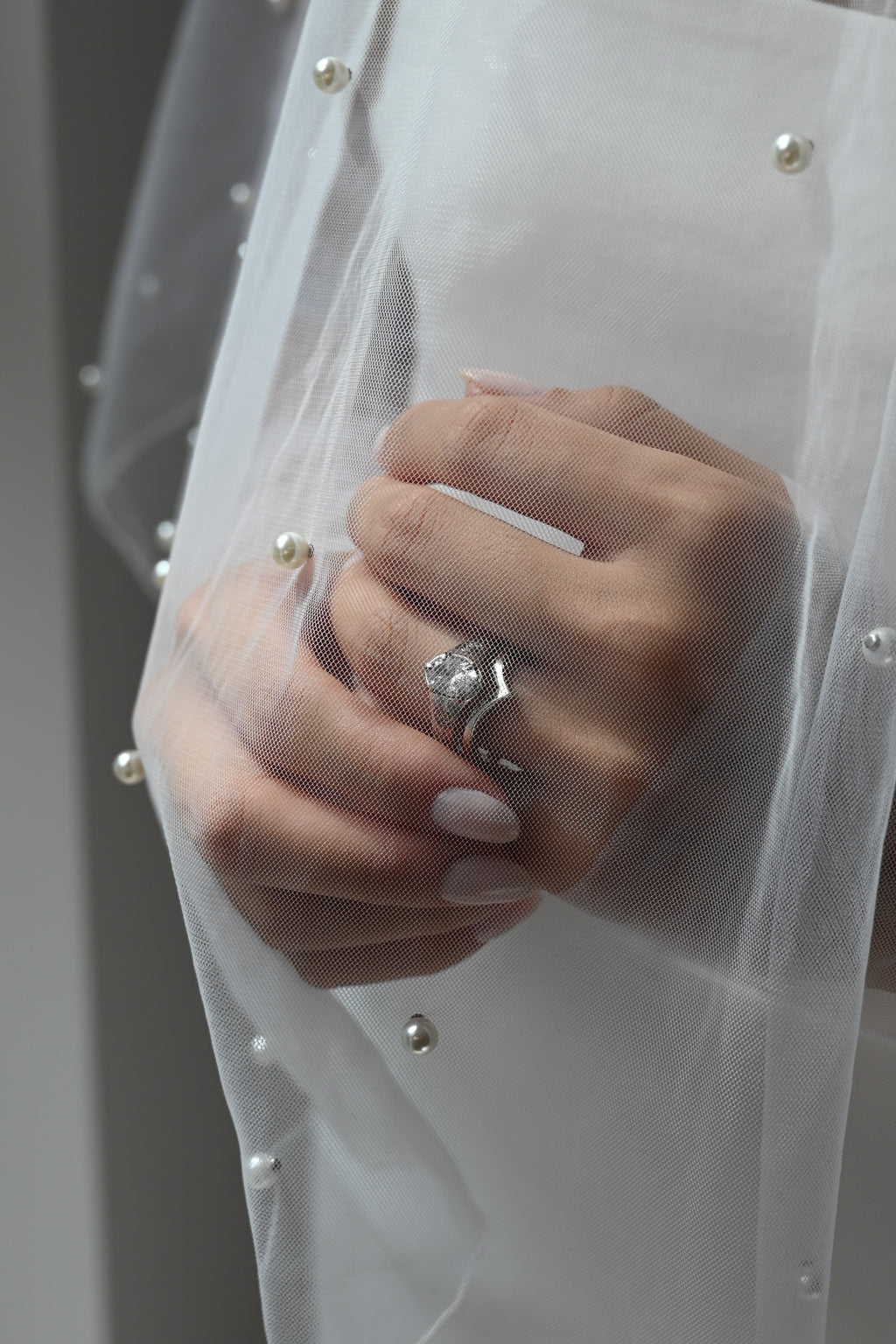 A close-up of a hand wearing a silver ring with a large clear marquise-cut gemstone, partially covered by a white pearl-trimmed sheer veil.