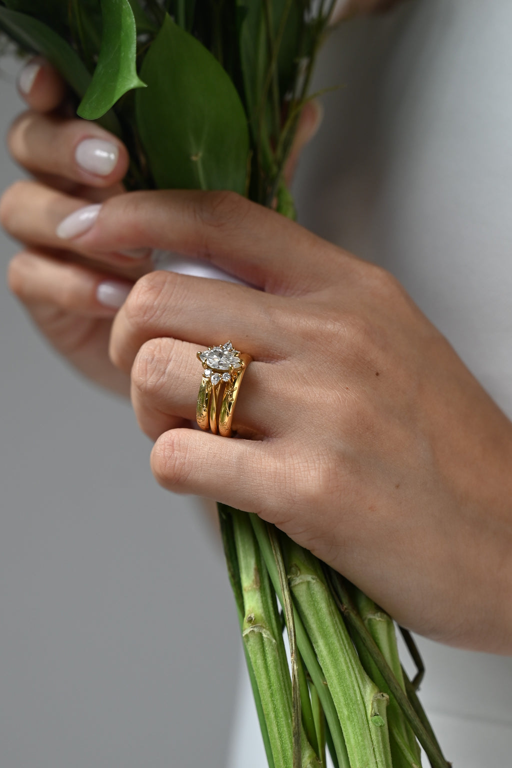 A close-up of a person's hands holding a green leafy bouquet, wearing a gold engagement ring with a round clear gemstone and a matching gold wedding band.