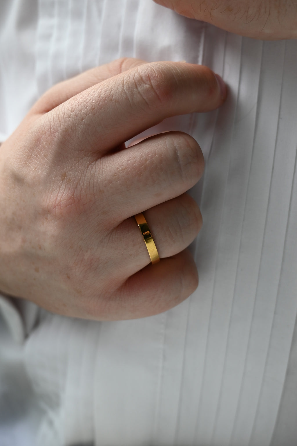 A close-up of a person's hand wearing a simple flat gold band ring, resting against a white pleated shirt.