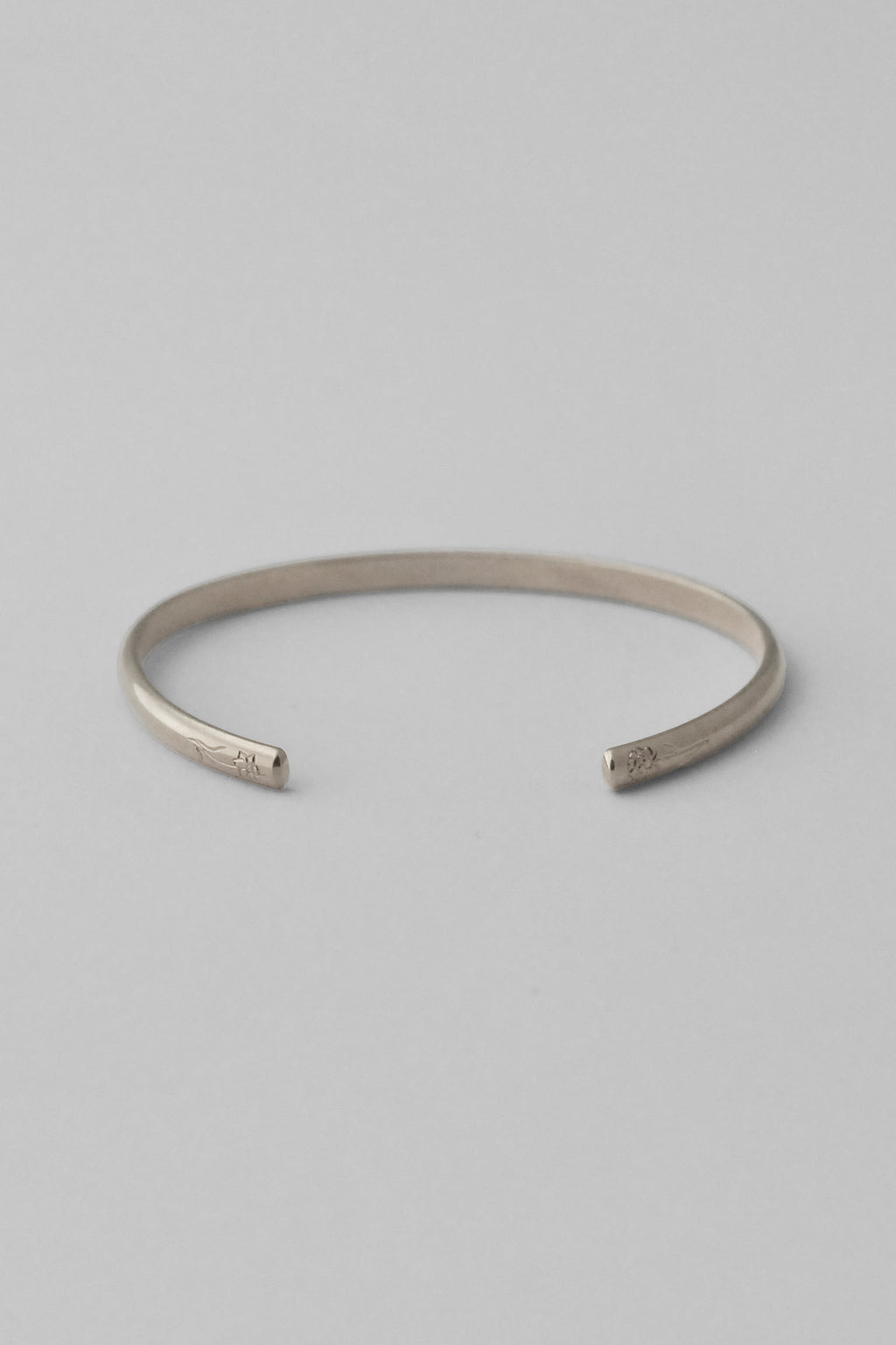 A minimalist sterling silver open cuff bangle bracelet with hand engraved flowers on each end, photographed on a clean, white background.

