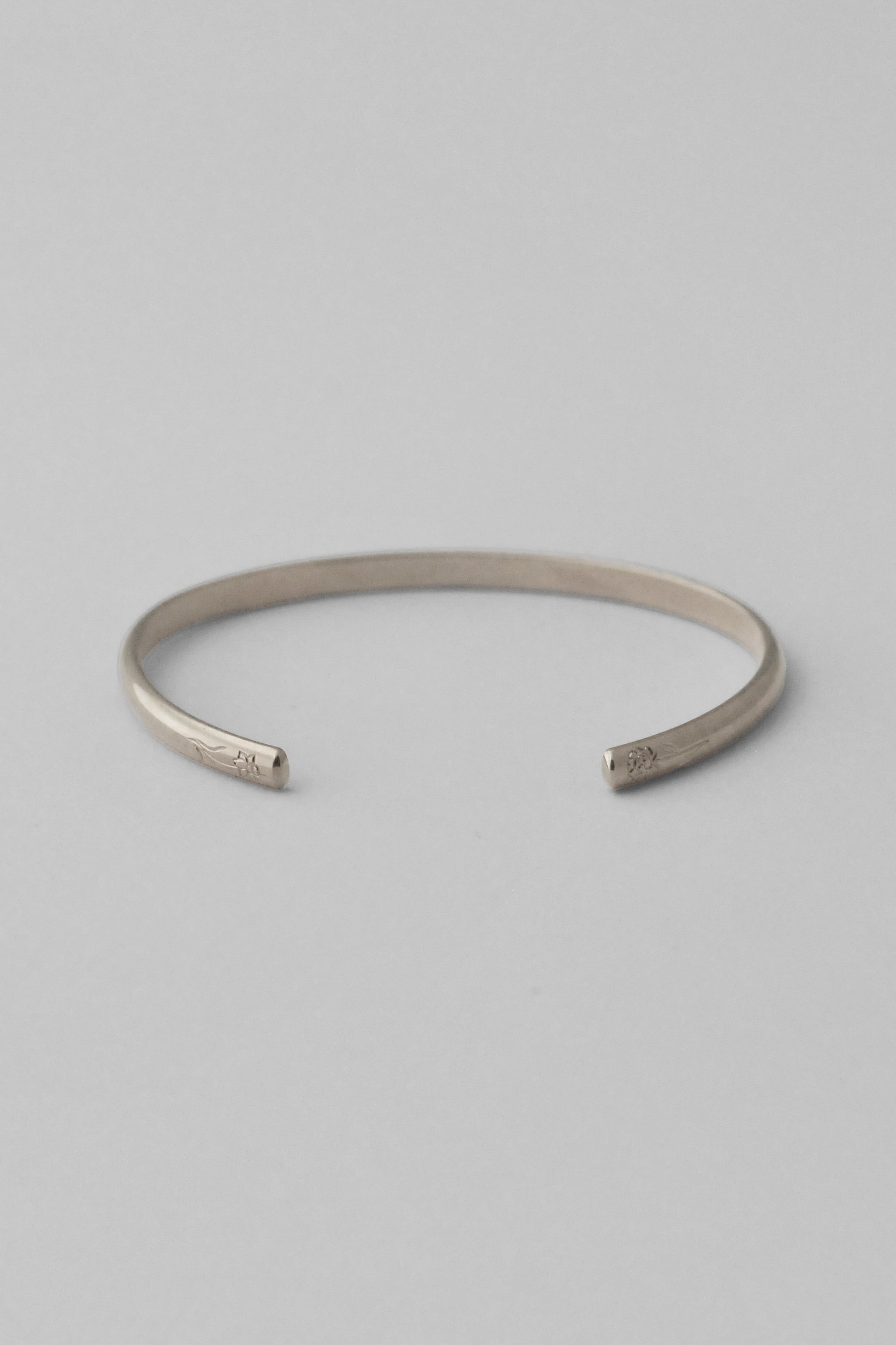 A minimalist sterling silver open cuff bangle bracelet with hand engraved flowers on each end, photographed on a clean, white background.
