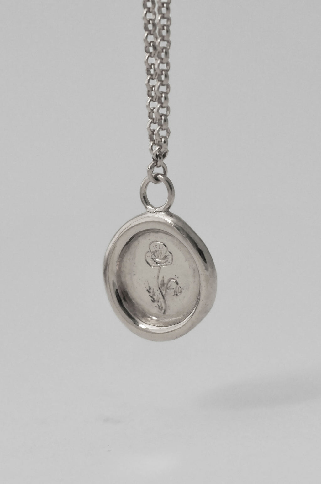 A silver necklace with a chunky organic wax-seal medallion pendant featuring a hand engraved birth flower floral engraving resting on a light white background.