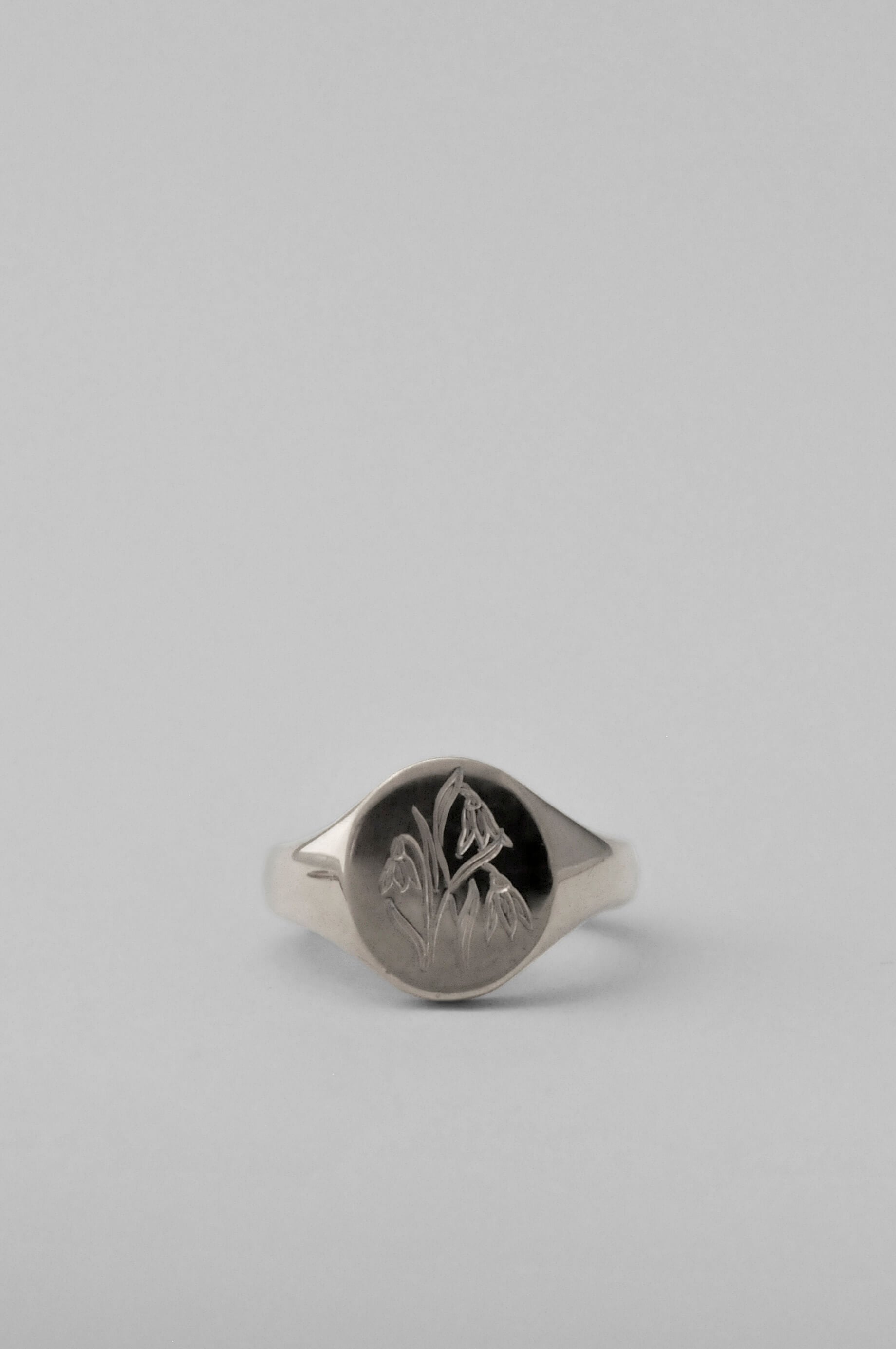 A silver signet ring with an engraved floral birth flower motif. Polished oval pinky signet ring, set against a solid light grey background.
