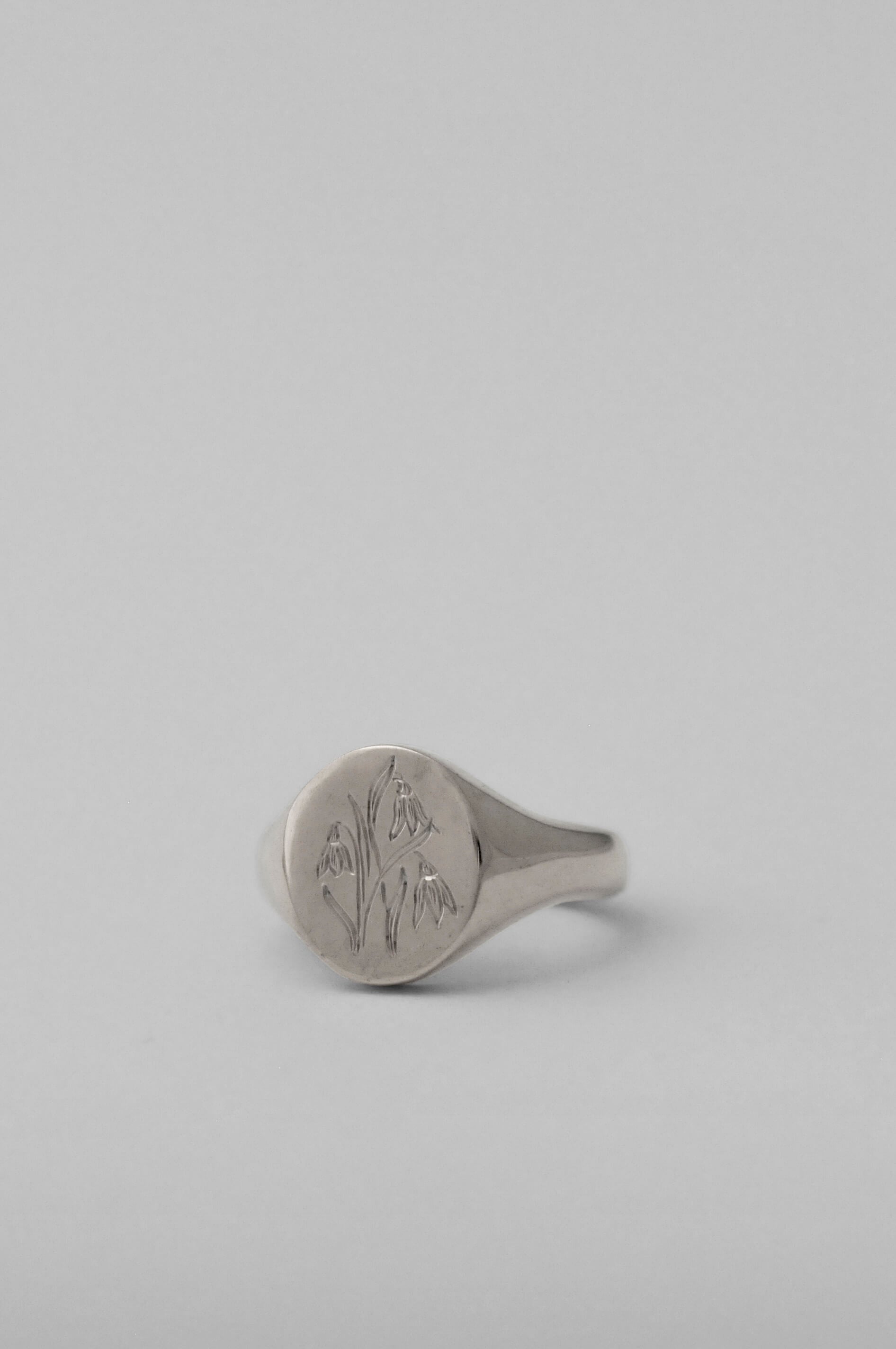 A silver signet ring with an engraved floral snowdrop flower motif. Polished oval pinky signet ring, set against a solid light grey background.